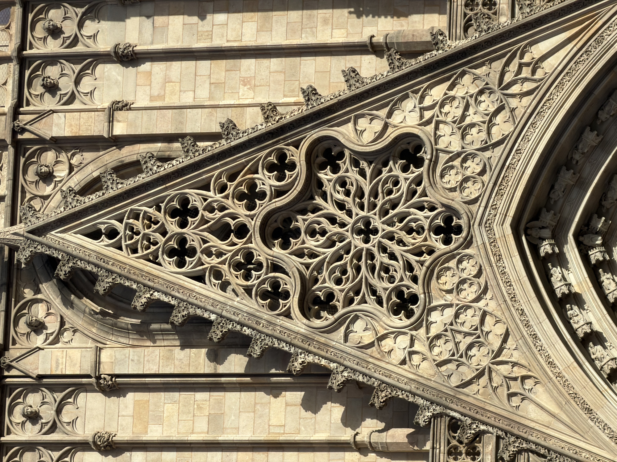The image shows the intricate Gothic facade of the Barcelona Cathedral, specifically a portion of the main entrance. The facade is constructed from light-colored stone blocks and features elaborate stonework, including pointed arches, tracery, and decorative carvings. Prominently featured is a large, triangular tympanum filled with detailed geometric and floral patterns, characteristic of Gothic architecture. Within the tympanum, a circular rose window with quatrefoil motifs is visible. Above this, rows of circular medallions contain carved human faces, some wearing head coverings. The stonework exhibits the warm tones of sandstone, illuminated by sunlight, casting shadows that highlight the depth and complexity of the ornamentation. The overall impression is one of grandeur and historical architectural artistry.