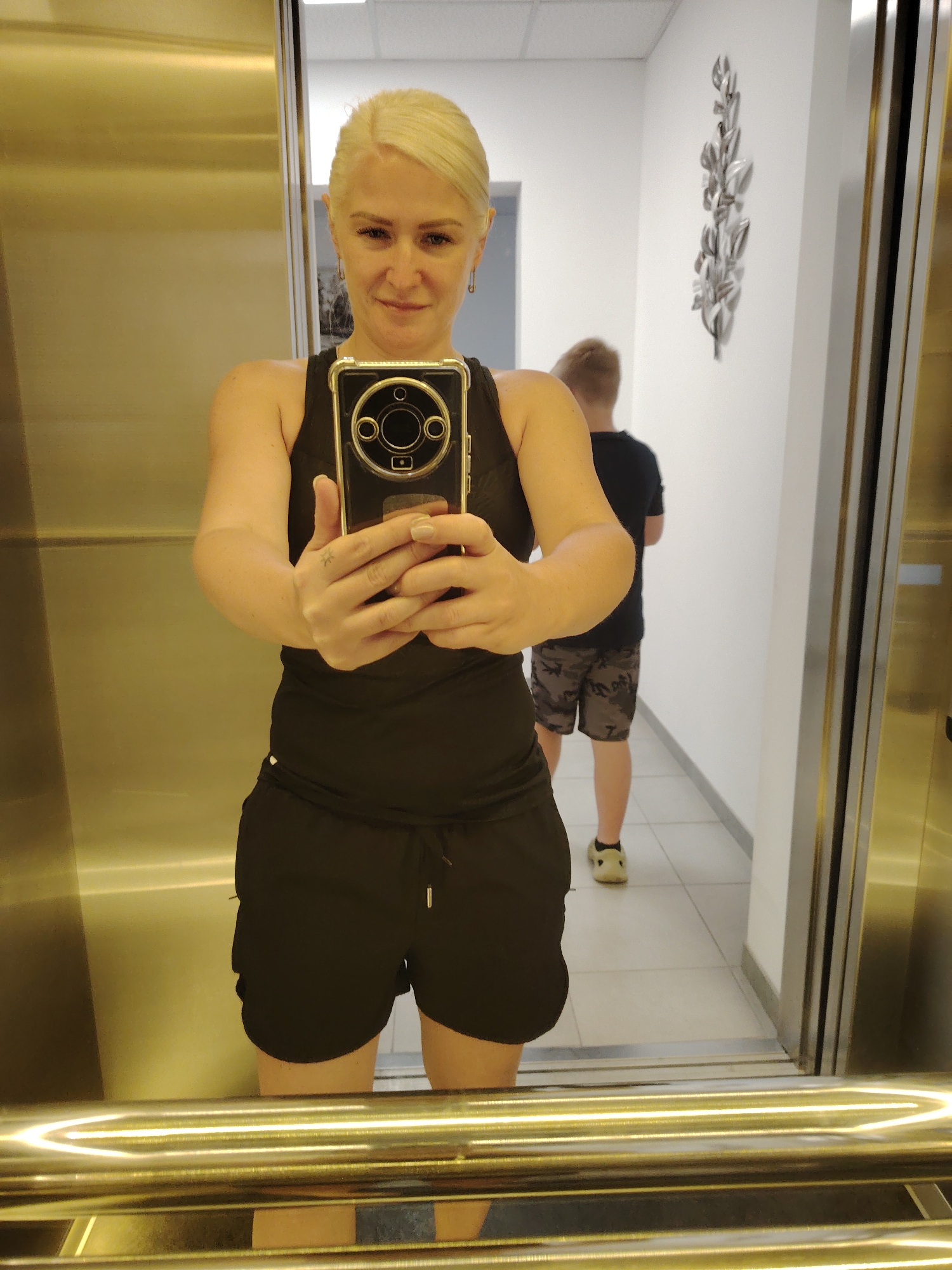 A blonde woman in a black tank top and black shorts takes a selfie in a mirrored elevator. She holds a smartphone in her right hand, its camera facing the mirror. A young boy in a black t-shirt and camouflage shorts stands in the background, facing away from the camera. The elevator walls are mirrored and metallic. A decorative silver leaf art piece hangs on the white wall in the background. The floor is tiled. The scene appears to be in Chaiky, Ukraine.
