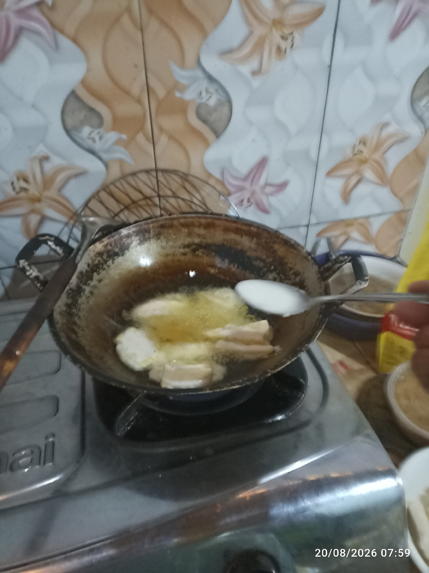 The image depicts a cooking scene, likely in a residential kitchen in Munjungan, Indonesia. In the foreground, a gas stove is visible, with a wok-like pan on top containing hot oil. Several pieces of food, appearing to be plantains or bananas, are being fried in the oil. A metal spoon is being used to add a white liquid, possibly batter or coconut milk, to the pan. The background shows a tiled wall with a floral pattern in beige and white. To the right of the pan, a yellow object and a white bowl containing a granular substance are partially visible. A metal rack is positioned behind the pan. The timestamp "20/08/2026 07:59" is overlaid on the bottom right of the image. The overall impression is one of home cooking in progress.