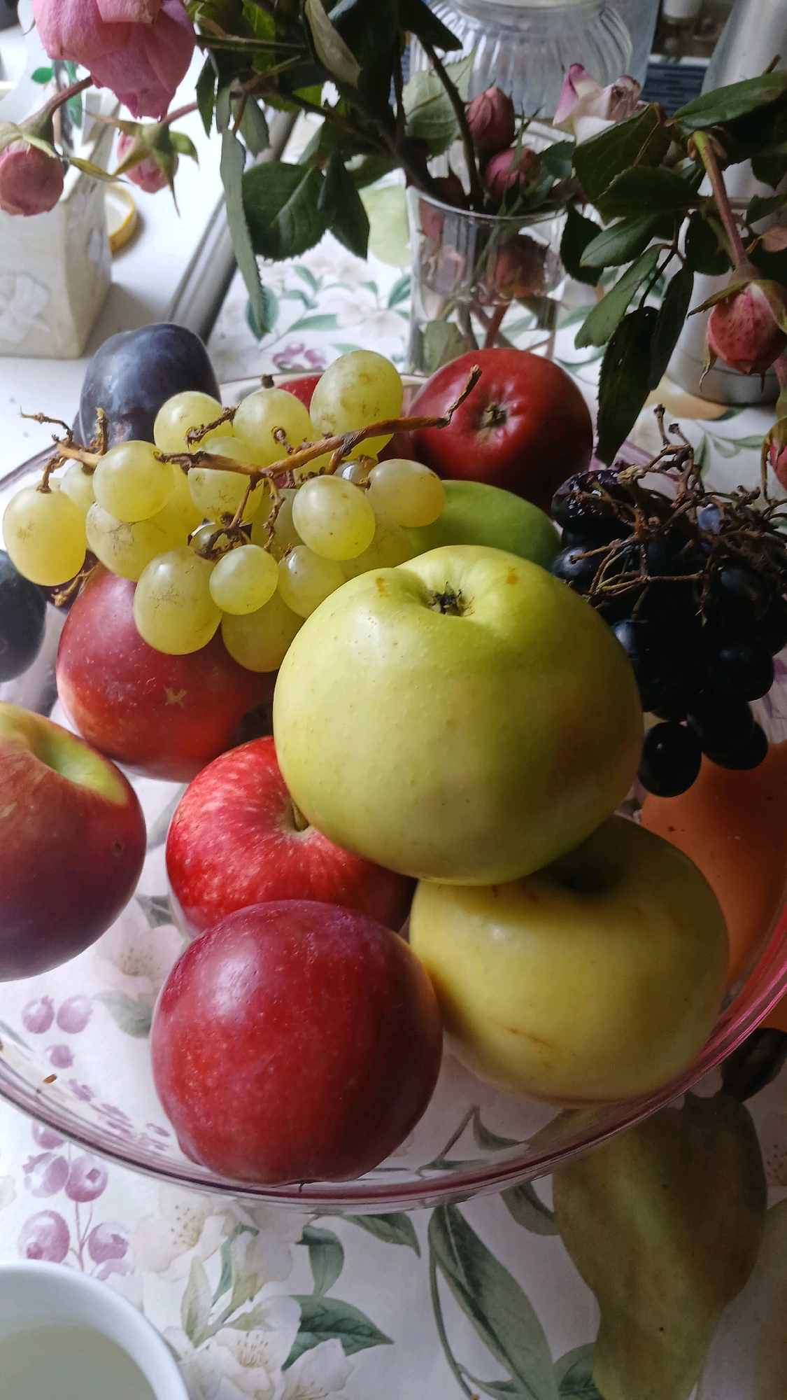 The image displays a still life composition of various fruits, including apples, grapes, and plums, arranged in a clear glass bowl. The fruits are a mix of red and green apples, green and dark purple grapes, and a dark purple plum. Some of the fruits appear to be of different varieties and sizes. In the background, a bouquet of pink roses and greenery is visible in a glass vase, suggesting a domestic setting. The surface beneath the bowl and vase is covered with a patterned tablecloth featuring floral designs. The lighting suggests an indoor environment, possibly a dining table or countertop. The image does not contain any information that would allow for its localization to Visaginas, Lithuania, or any specific city. There are no people, actions, or interactions depicted.