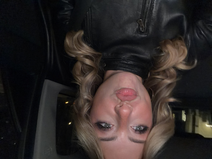 A young female with long, wavy blonde hair and light-colored eyes is captured in a close-up image, seated inside a vehicle at night in the city of Paris, France. She has prominent dark eyeliner, a reddish hue on her nose and cheeks, and her full lips are pursed in a 'kissing' or 'pout' expression. She is wearing a black leather jacket with a visible front zipper and a high collar, and also has small gold-colored hoop earrings. The background, visible through the vehicle's windows, shows blurred yellow and orange city lights and indistinct, dimly lit building structures, indicating an urban environment after dark.