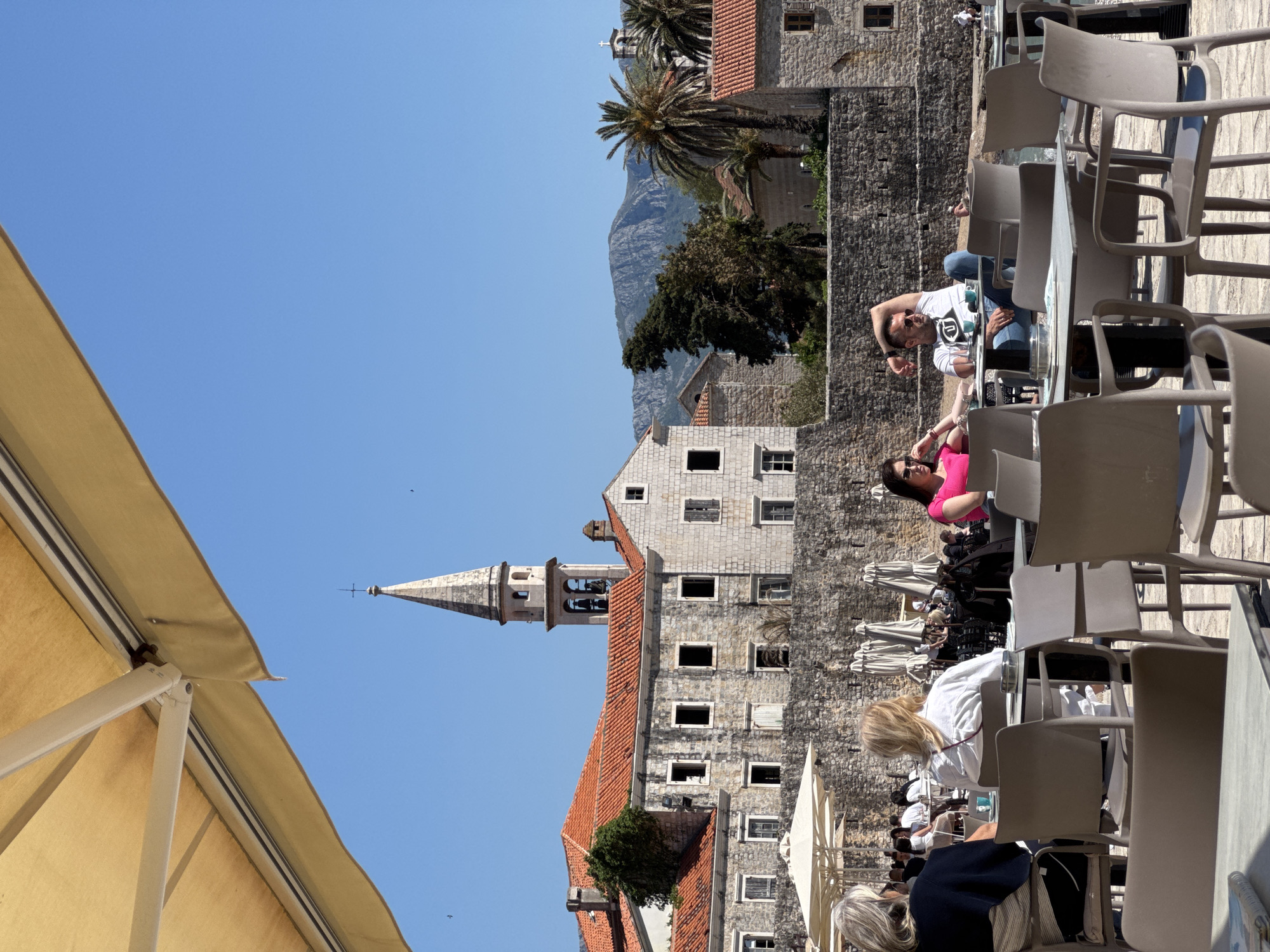 The image shows a sunny, outdoor scene in Budva, Montenegro. A tall, stone church with a pointed steeple is visible in the background, with a building attached to it. The building has a red-tiled roof and several windows. In the distance, there's a mountain range. Palm trees are also present in the background.
In the foreground, there's an outdoor café or restaurant area with numerous empty chairs and tables. Several people are sitting at some of the tables, including a couple visible at one of the tables; one is wearing a white shirt and the other is wearing a pink top. Some people are partially obscured by chairs and tables. There's a yellow awning partially visible at the edge of the frame.