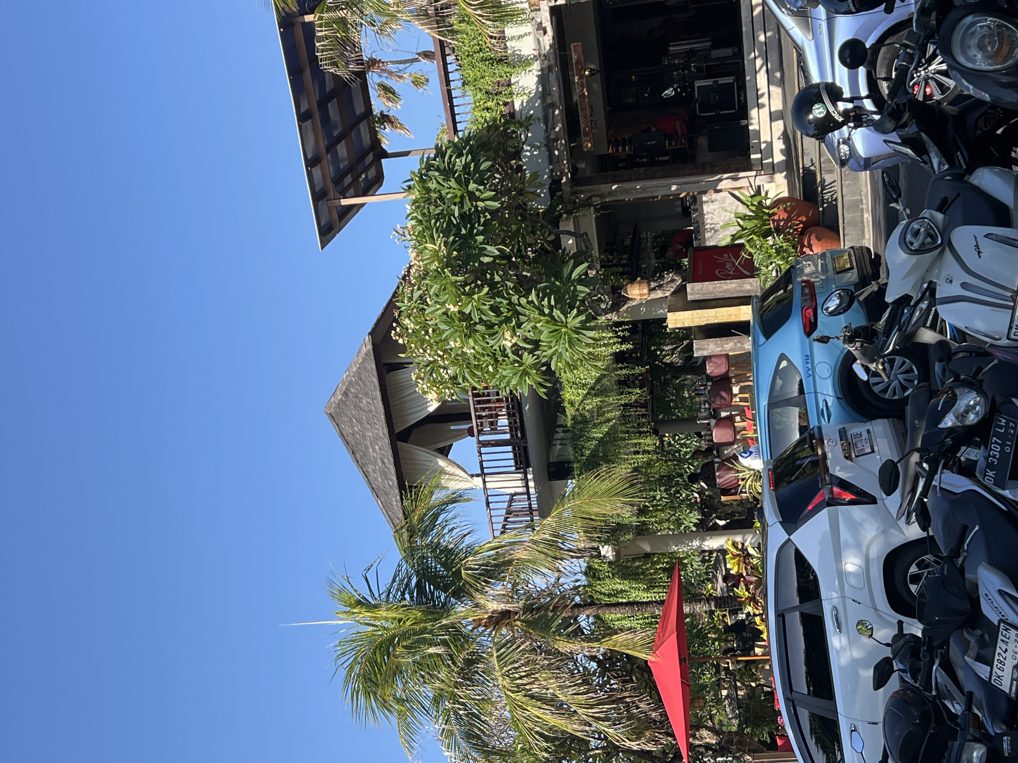 In Kuta, Indonesia, a clear blue sky stretches above a multi-story building with a thatched roof and balconies adorned with lush greenery. A large palm tree stands to the left, its fronds reaching towards the sky, with a vibrant red umbrella positioned near its base. In front of the building, a row of parked vehicles includes a white SUV and a light blue car, with several scooters parked in the foreground. People are visible in the background, seated at tables under the building's covered areas, suggesting an outdoor dining or seating arrangement. The scene captures a sunny day in a tropical urban environment.