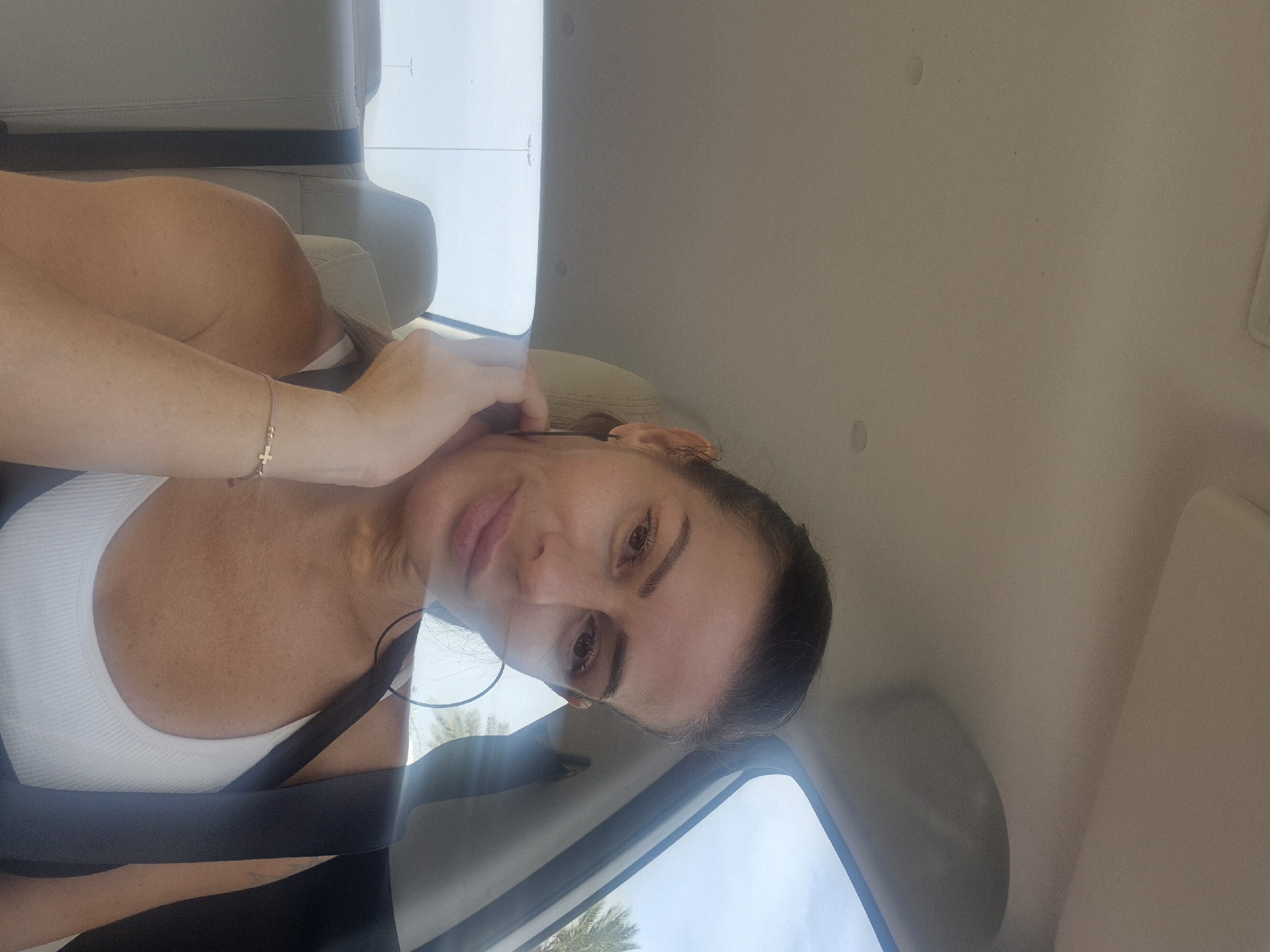 A woman with dark hair is seated in a car, wearing a white tank top and a black seatbelt. She has a bracelet on her left wrist and a hoop earring in her ear. She is holding her hair near her ear with her hand. The interior of the car is beige, and outside the window, palm trees and a blue sky are visible, consistent with the city of Dubai, United Arab Emirates.