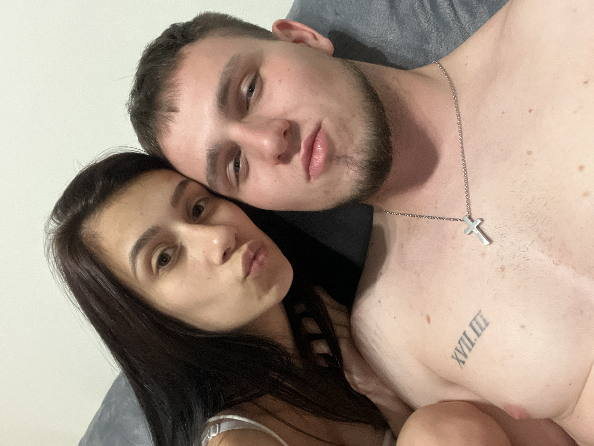 A selfie shows a man and a woman lying closely together. The man has fair skin, short brown hair, a beard, and a silver necklace with a cross pendant. A tattoo in Roman numerals is visible on his chest. The woman, who rests her head against his, has long, dark hair and fair skin. Both subjects are pursing their lips. The background includes a gray fabric surface and a white wall. The location is not specified in the image.
