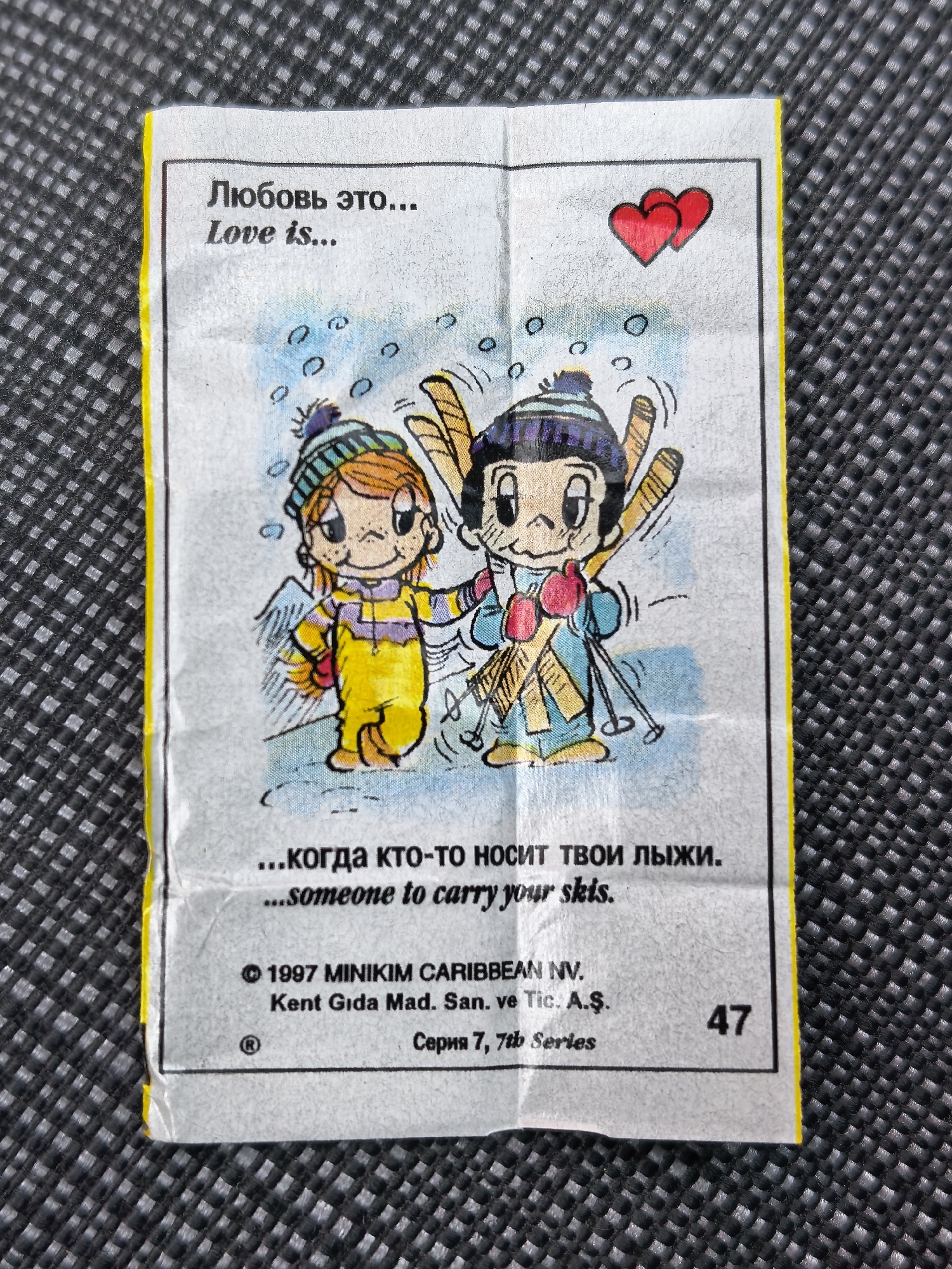 The image is a close-up, overhead shot of a small, folded paper insert, likely from a product. The paper is textured and slightly creased, with a bright yellow border around the perimeter. The center of the paper features a cartoon illustration of two children in winter clothing, holding skis. Above the illustration, text is printed in Russian and English. The Russian text reads "Любовь это..." and the English text reads "Love is...". Below the illustration, there is more text, with Russian reading "...когда кто-то носит твои лыжи." and English reading "...someone to carry your skis." At the bottom of the paper, copyright information is visible: "© 1997 MINIKIM CARIBBEAN NV. Kent Gida Mad. San. ve Tic. A.Ş." and a serial number "47". The background of the image is a dark, textured fabric, possibly a tablecloth or a surface with a raised, grid-like pattern. There is no indication of the city of Elektrostal, Russia, in the image or any contextual information provided.
