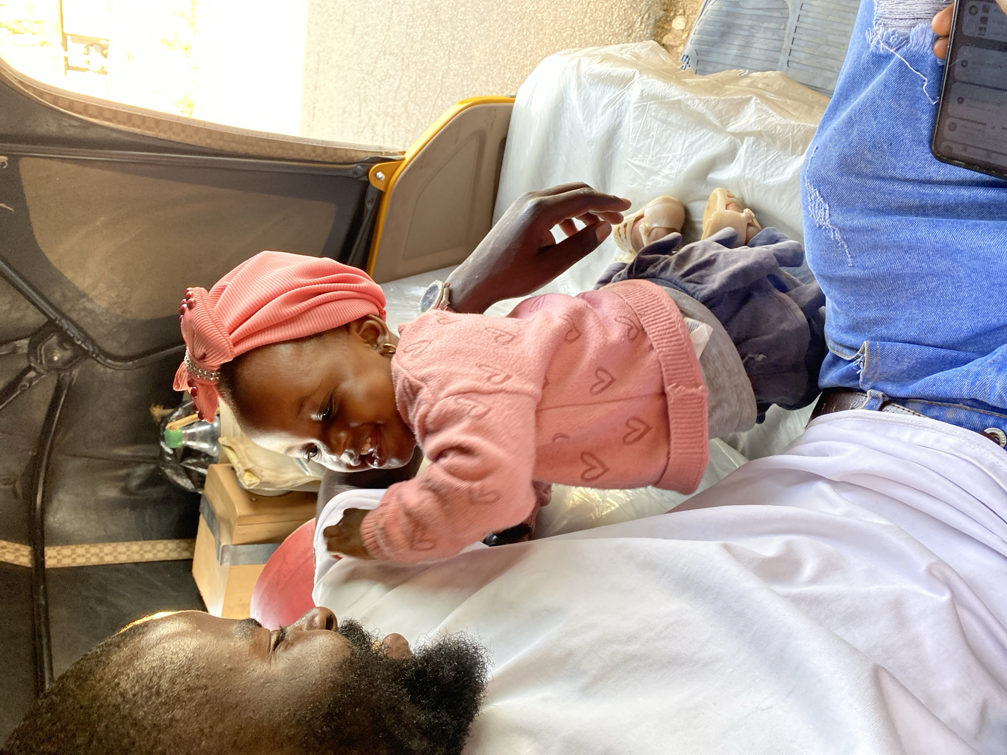 An overhead shot captures an intimate moment inside what appears to be a three-wheeled auto-rickshaw (Keke Napep) in Vom, Nigeria. The main subjects are a man and a young child, positioned for a close interaction.
The child, wearing a pink ribbed headwrap adorned with beads and a light pink long-sleeved sweater with small heart patterns, is seen leaning forward and looking down with a gentle smile. Her head is angled towards the man, and her small hands are resting near his chest. She also wears sandals on her feet.
The man, with a dark, full beard, is positioned in the lower left of the frame, lying or reclining. He is looking up towards the child with a soft, engaged expression. He is dressed in white fabric, possibly a traditional garment or a simple sheet.
In the upper right, a portion of another person is visible, wearing blue ripped jeans and holding a smartphone, suggesting they might be a passenger or driver. A dark-skinned arm with a wristwatch is also visible near the child. The interior of the vehicle features dark seating, a light-colored protective covering or sheet, and a yellow-framed structure typical of such vehicles. A wooden box and a plastic bottle are seen in the background on the left.
The bright natural light indicates it is daytime. The overall mood is one of warmth, affection, and connection between the child and the man. No discernible text is visible on any objects or screens.