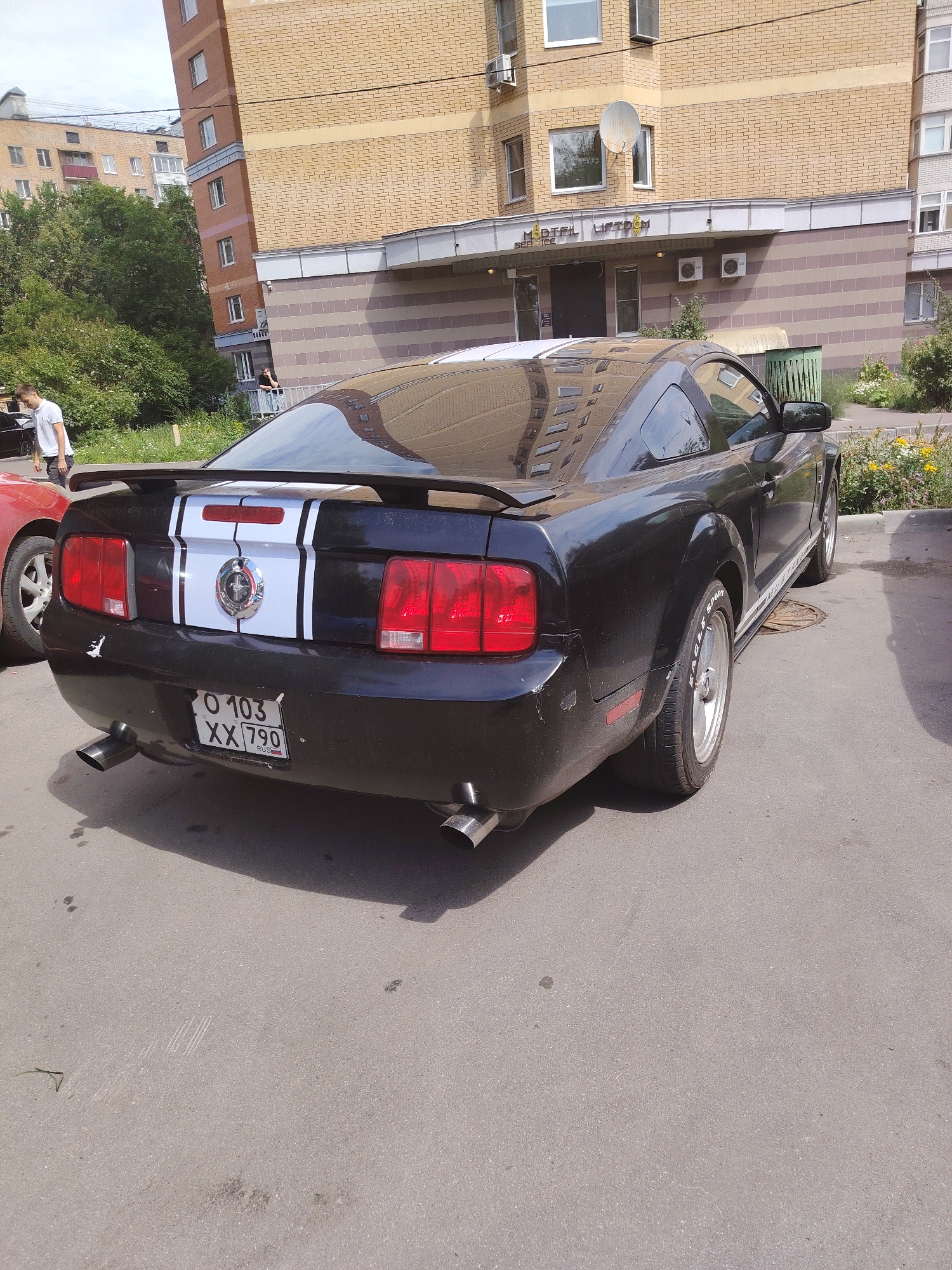 A black Ford Mustang is parked on an asphalt surface in front of a brick building. The car has white racing stripes on its trunk and a chrome emblem on the rear. Its license plate is visible and reads "O 103 XX 790 RUS". A red car is partially visible to the left of the Mustang. A man in a white t-shirt and grey shorts stands on the sidewalk to the left of the cars, facing away from the camera. The building behind the cars has a tan brick facade with a brown and grey striped lower section. A satellite dish is mounted on the side of the building. The image appears to have been taken in Moscow, Russia, given the license plate format and the architectural style of the building.