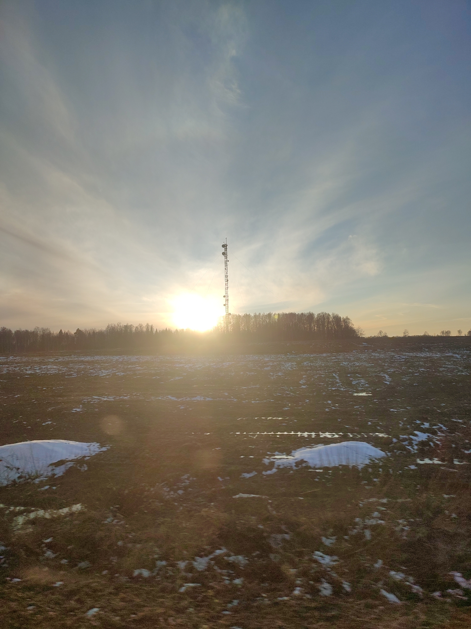 The image shows a rural landscape under a bright, slightly hazy sky with cirrus clouds. In the foreground is a field with patches of melting snow and water. A line of trees stretches across the middle ground, with a telecommunications tower rising prominently behind them. The sun is positioned behind the trees, creating a strong glare. The location is Unknown, Unknown.