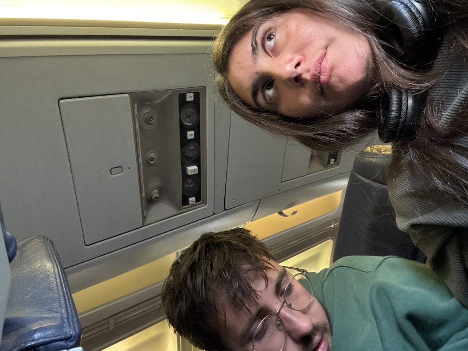 The scene is an interior view of an aircraft cabin, located in the city of Tzafria, Israel. Two individuals occupy seats in the cabin. On the left, a male with dark hair and gold-rimmed glasses is depicted with his head tilted to his right, wearing a green hooded garment, and appearing to be asleep or resting. On the right, a female with long dark hair and brown eyes is looking upwards and to her right. Black over-ear headphones are resting around her neck over a grey, ribbed top. The overhead cabin panel is visible, featuring air vents, reading lights, and call buttons. A partially visible blue-grey overhead bin is in the upper left. Yellowish light from a window or cabin lighting illuminates the right side of the frame and the background. The seating appears to be dark-colored.