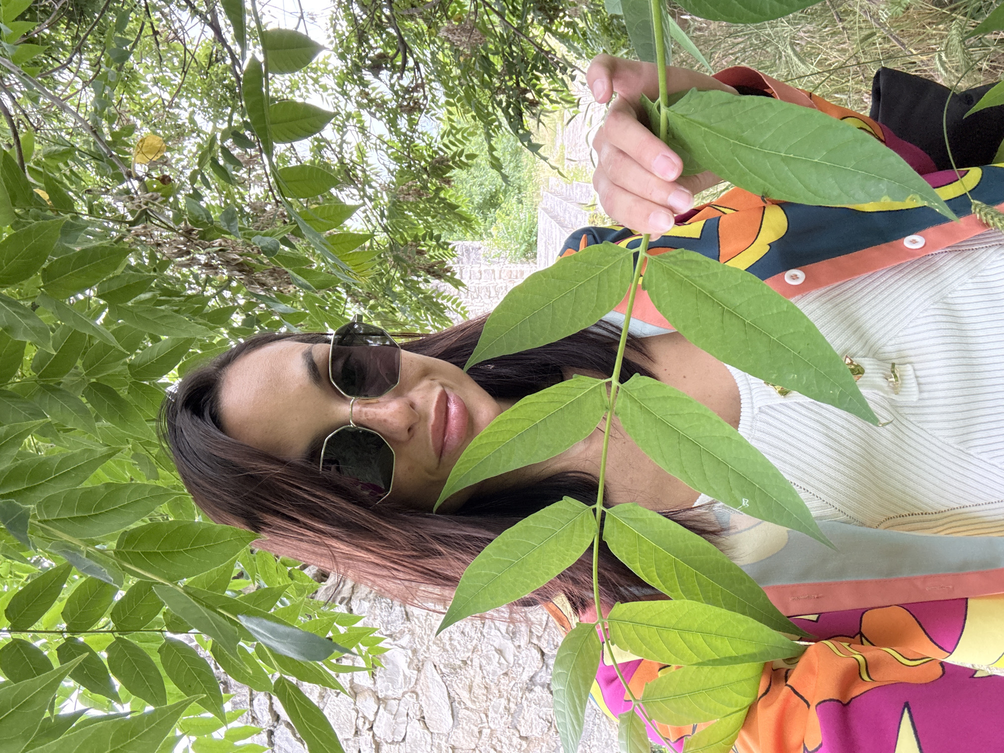 A woman with dark hair, wearing sunglasses and a colorful patterned jacket over a ribbed white top, holds a branch with green leaves in front of her face. She is positioned outdoors, with a stone wall and greenery in the background, possibly in Pleven, Bulgaria, given the context.