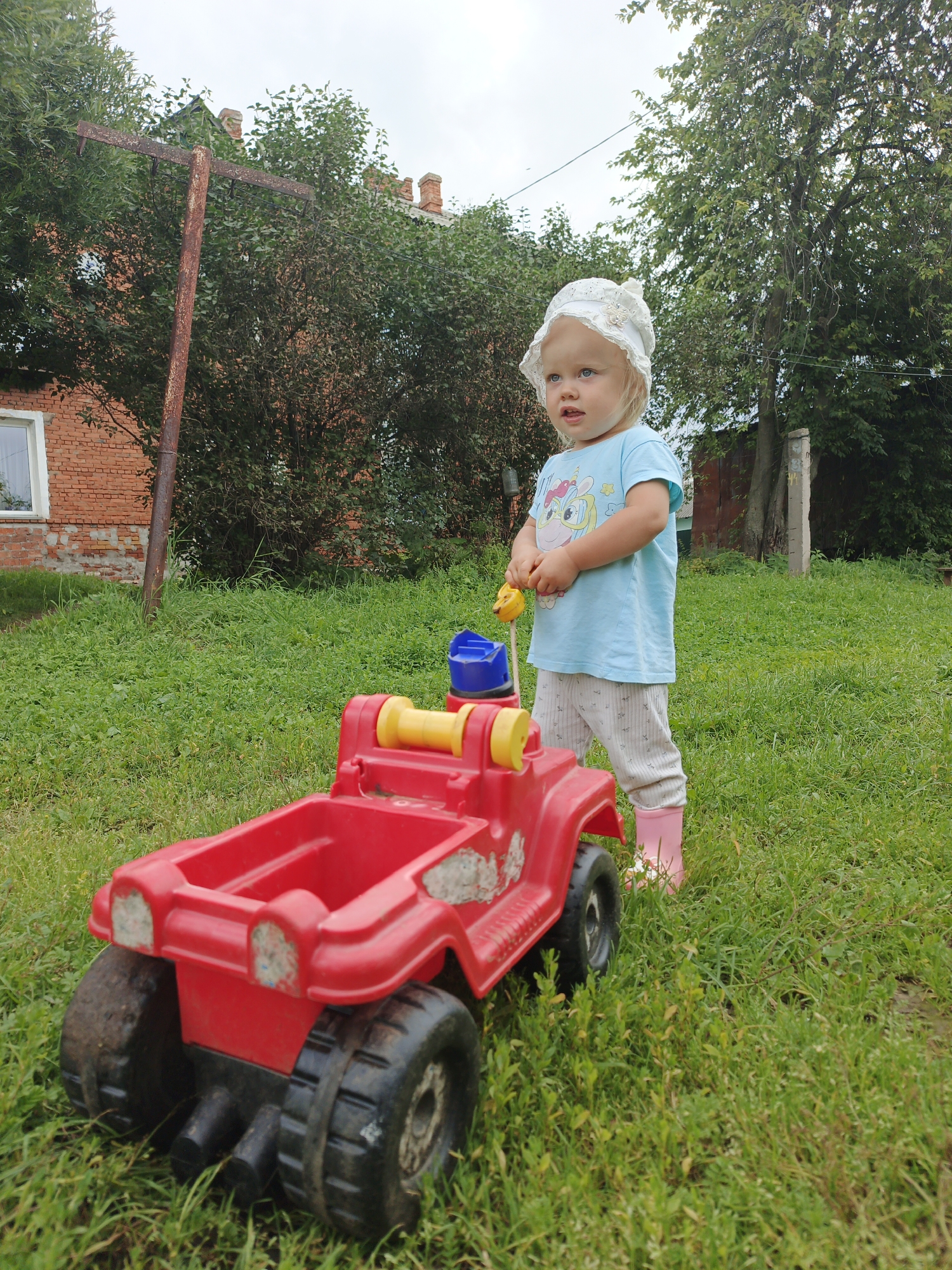 A young Caucasian child, likely a toddler, stands in a grassy yard in Medvedyovka, Russia, next to a large red toy jeep. The child is wearing a light blue t-shirt with a unicorn graphic, light-colored pants with a subtle pattern, and pink rubber boots. A white sun hat with lace trim is on their head. They are holding a yellow toy in their right hand, and their left hand rests on the toy jeep's steering wheel. The toy jeep is red with black oversized wheels and yellow and blue accents. In the background, a brick building with a white-framed window is visible, partially obscured by green trees. A rusty metal pole with a horizontal bar is also present in the yard, with power lines running across it. The sky is overcast.