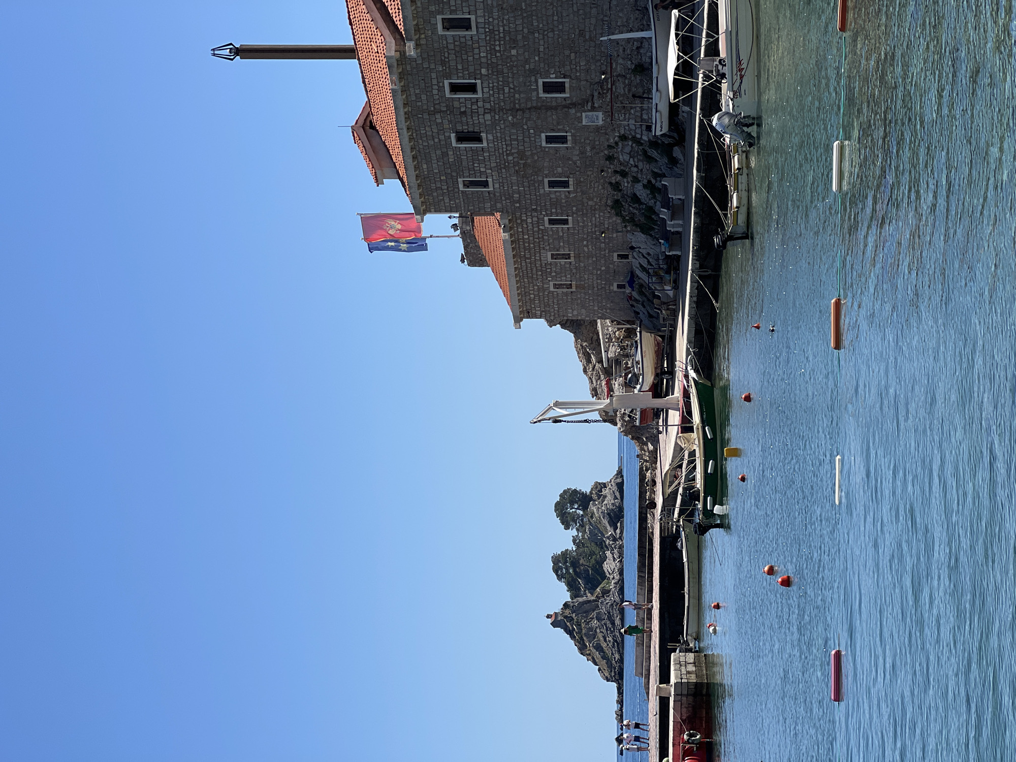 This image captures a vibrant, sunny coastal scene in what appears to be Petrovac centar, Montenegro. The clear, deep blue sky fills the upper half of the frame, indicating excellent weather conditions. Below, calm, blue-green water stretches across, reflecting the sunlight and showing gentle ripples.
On the left, a sturdy stone building with a red-tiled roof stands by the water. Two flags are prominently displayed on poles near this structure: a red flag with a golden emblem, which is the national flag of Montenegro, and a blue flag adorned with white stars, likely representing the European Union. A tall, dark pole with an ornate top extends upwards from the building's roofline.
Along the waterfront, a quay or promenade is visible, where several boats are moored. One white boat with an outboard motor is particularly noticeable. The water surface is marked by numerous buoys of various colors—orange, white, red, and yellow—interconnected by ropes, suggesting a designated area for vessels or swimmers. Further along the coastline, rugged rocky outcrops with sparse green vegetation extend into the sea. A white crane structure is also present on the quay.
A few indistinct figures of people can be seen on the promenade, enjoying the pleasant weather and coastal ambiance. The bright sunlight and absence of clouds suggest it is midday or early afternoon. The scene is peaceful and picturesque, characteristic of a Mediterranean port town. No discernible text is visible within the image.