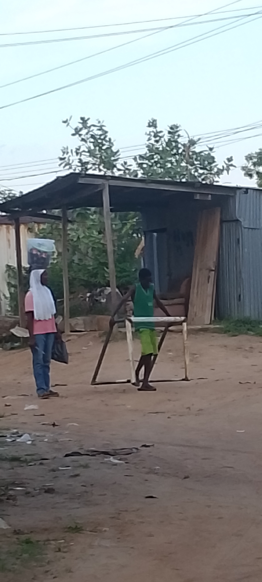 The image shows a street scene in Adankolo, Nigeria. A woman wearing a white headscarf, pink shirt, and jeans is standing to the left of a makeshift structure with a corrugated metal roof. She is carrying a dark-colored bag. A young man in a green tank top and green shorts is to the right, holding onto a small, possibly homemade, goalpost frame. The ground is dirt. Behind the structure are trees and a corrugated metal building. Trash is scattered on the ground in the foreground. Overhead are power lines.