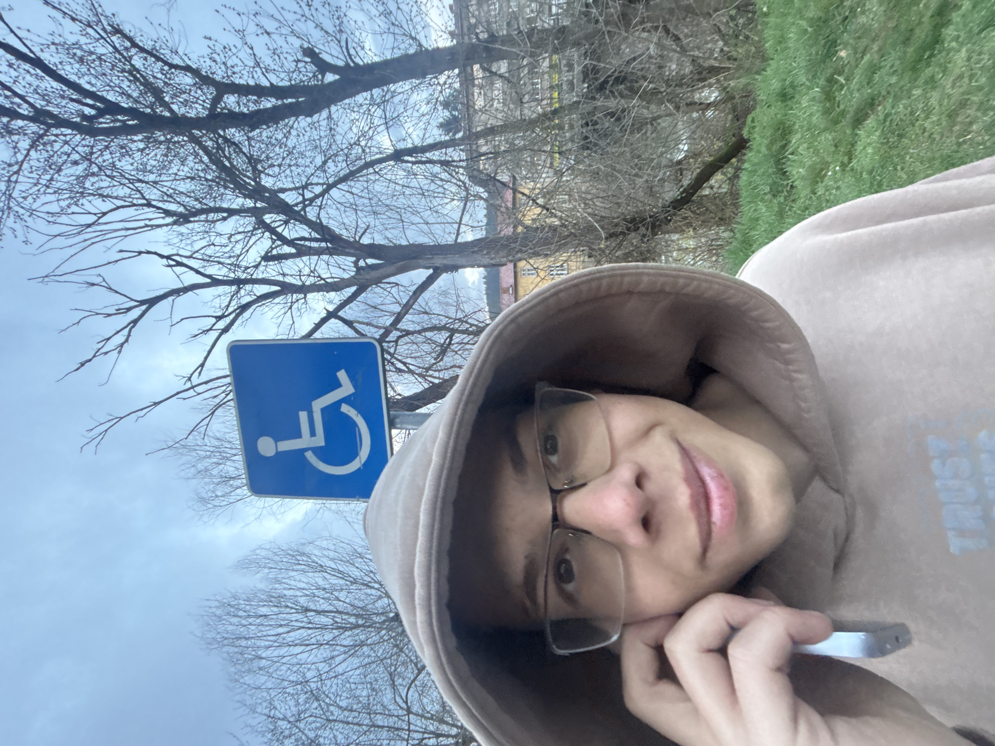 A close-up selfie, taken outdoors, presents a person with fair skin wearing glasses and a light brown hooded sweatshirt. The person is holding a small, metallic object near their ear. Behind the person and to the left, a blue and white handicapped parking sign is visible. Bare trees frame the background, partially obscuring a building with a yellow wall and red accents. In the upper right corner, a patch of green grass is visible. The sky is overcast. The location is the city of Troyan, Bulgaria.