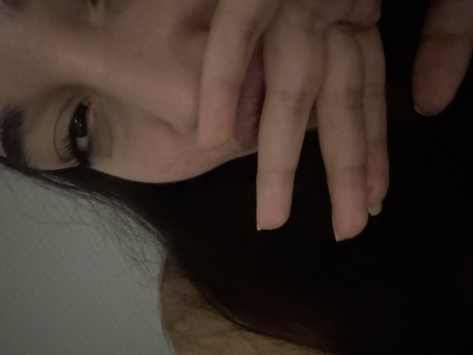 A selfie-style image features a person's face and hand, partially obscured by darkness. The face, slightly tilted, shows an eye with dark lashes and part of the nose. The hand, with fingers partially covering the mouth, appears close to the camera. Long, dark hair frames the face. The background is dimly lit, making details indistinct.