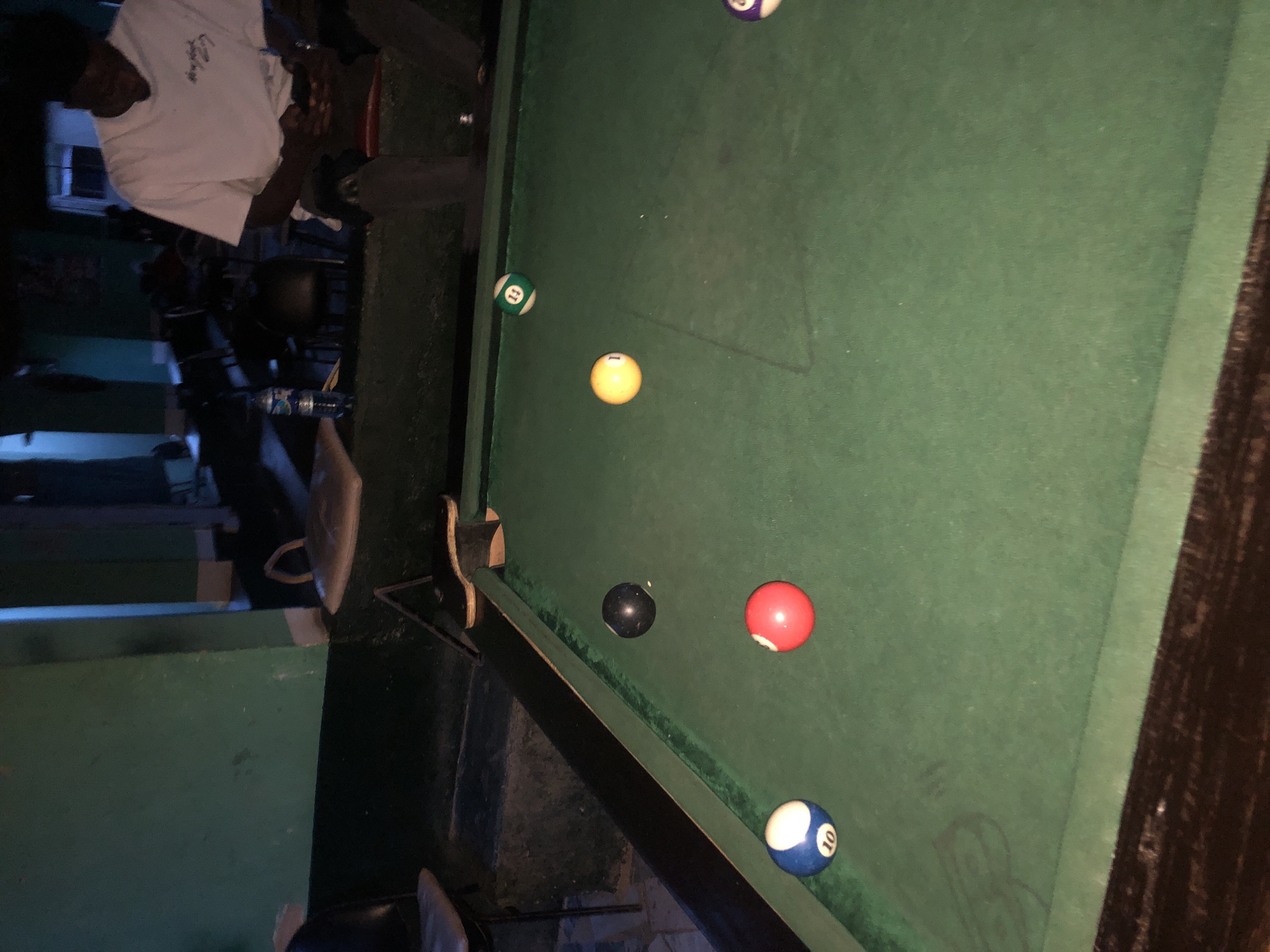 The image depicts an indoor scene within an establishment in Unknown, Unknown, characterized by low light conditions. The foreground and mid-ground are dominated by a green-felted pool table with dark rails. Six pool balls are scattered across the table: a blue and white striped 10-ball near the bottom left cushion, a solid red ball (number indiscernible) towards the bottom center, a solid black 8-ball near the top left cushion, a solid yellow 1-ball in the upper-mid section, a green and white striped 11-ball near the top center cushion, and a partial view of a purple and white striped ball (number indiscernible) on the far right edge.
In the background, positioned to the right of the pool table, a male individual is seated on a dark chair with a reddish cushion. He wears a white t-shirt with dark script on the chest and dark trousers. His gaze is directed downwards at a small electronic device held in his hands, likely a mobile phone. To his left, on a raised dark platform behind the pool table, a plastic water bottle with a blue cap and a partially visible "AQUA" label is present. Also on this platform, further left, is a light-colored, possibly fabric, object with handles, resembling a cushion or bag. In the deeper background, various dark tables and chairs are visible, out of focus. The walls in the background are painted a dark green, exhibiting some visible wear and tear. The overall lighting is dim, casting shadows and obscuring precise details in the periphery.