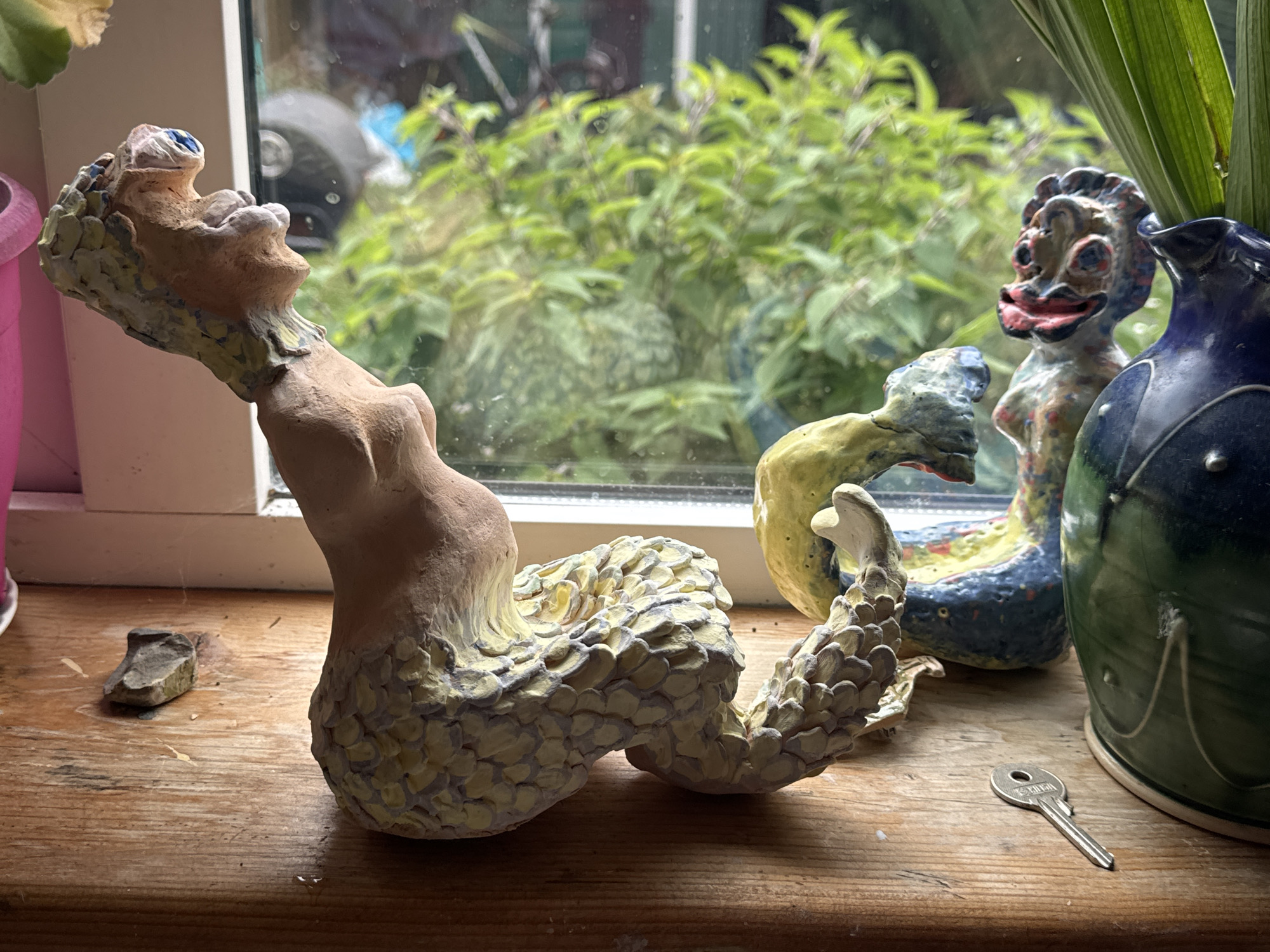The image depicts a windowsill in what appears to be an artist's studio or home, set against a blurred outdoor view of lush green foliage. The windowsill is made of weathered, light brown wood and is cluttered with various objects.
In the foreground, prominently placed, is a light brown ceramic sculpture of a creature with a human-like upper torso and a scaly, coiled tail. The creature's head is tilted back, with its mouth open, revealing a pink interior and a blue eye. The scales are painted in pale yellow and grey. To the left of this sculpture, a small, dark grey, irregularly shaped stone rests on the windowsill.
Further back on the windowsill, another ceramic sculpture is visible. This one has a more distinct humanoid form with a wide, smiling mouth, red lips, and a blueish-grey head with red and yellow accents. It appears to be a smaller, more stylized figure than the first.
To the right of the second sculpture, a large, dark green ceramic vase with a blue rim is present. A tall, green leaf emerges from the vase, partially obscuring the view of the second sculpture.
In the bottom right corner of the image, a small, silver key lies on the wooden windowsill, its teeth facing upwards.
The background, seen through a window, shows a dense, out-of-focus arrangement of green leaves and branches, suggesting an outdoor garden or natural setting. The window frame is white.
There are no people or overt actions depicted. The scene is static, presenting a collection of artistic creations and everyday objects in a natural light setting. The overall impression is one of a creative space, possibly a windowsill in a residential or studio environment in Waterford, Ireland, given the prompt's specific location request.