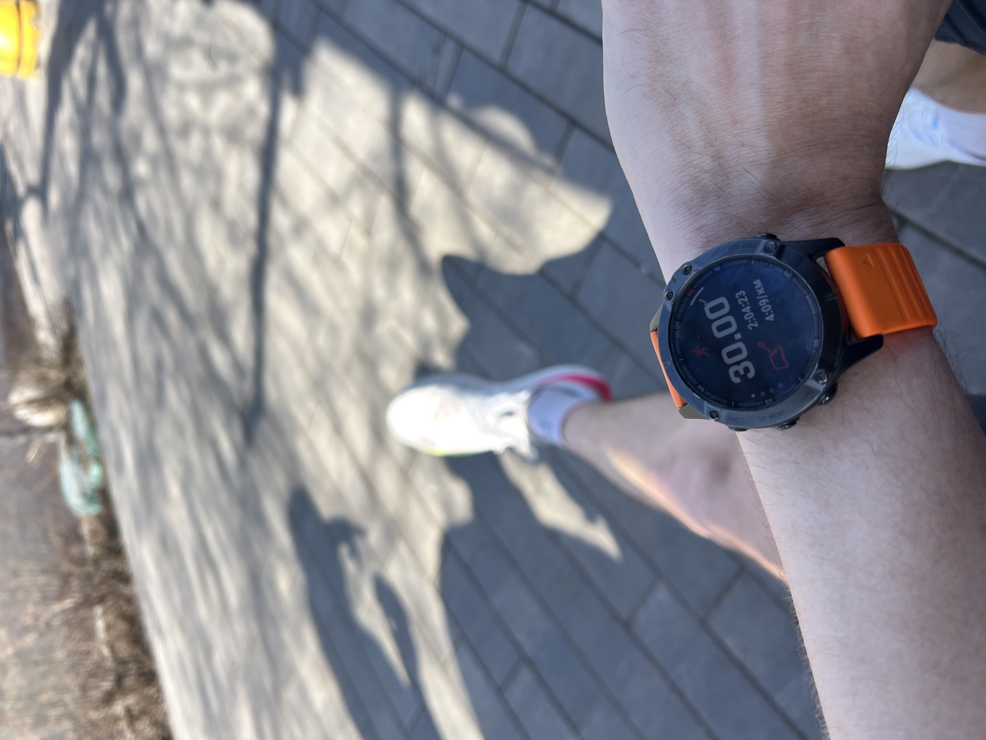 A person is wearing a black Garmin watch with an orange band on their left wrist. The watch face displays a time of 30.00, a duration of 2:04:23, and a pace of 4:09/km. The person is wearing a white athletic shoe with a pink accent. The person is standing on a paved surface, with shadows of trees and other objects visible. The location is not specified and cannot be inferred from the given details.