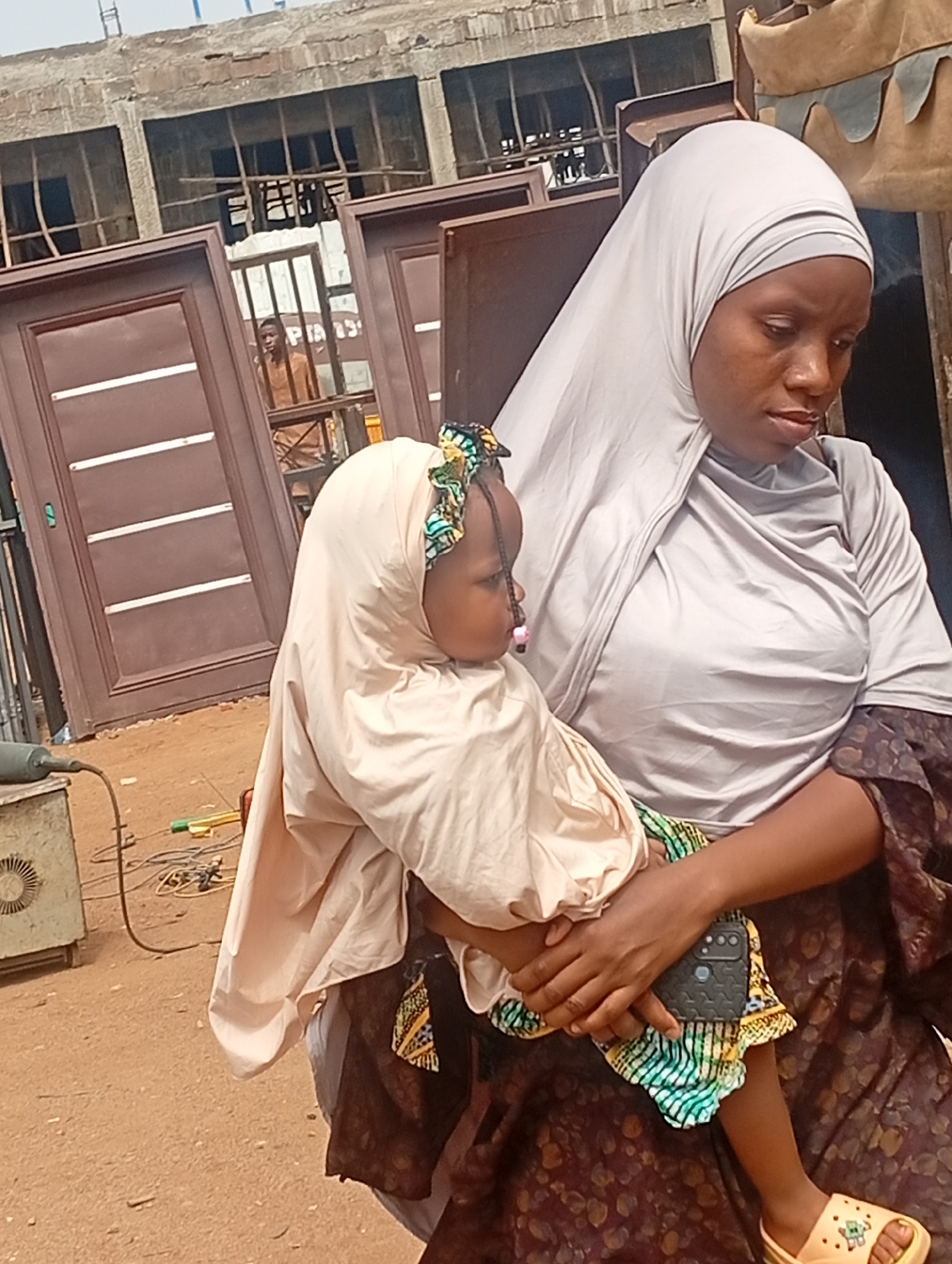 The image shows a woman wearing a light grey hijab and a dark brown patterned dress, holding a young child in her arms. The child is wearing a light beige hijab with a patterned headband and yellow sandals. The woman is holding a black smartphone in her left hand. In the background are brown metal doors, a man standing behind a metal gate, and an unfinished building. To the left of the woman, there is a piece of machinery with electrical cables on the ground. The ground appears to be a reddish-brown dirt surface. The image was taken in Jos, Nigeria.