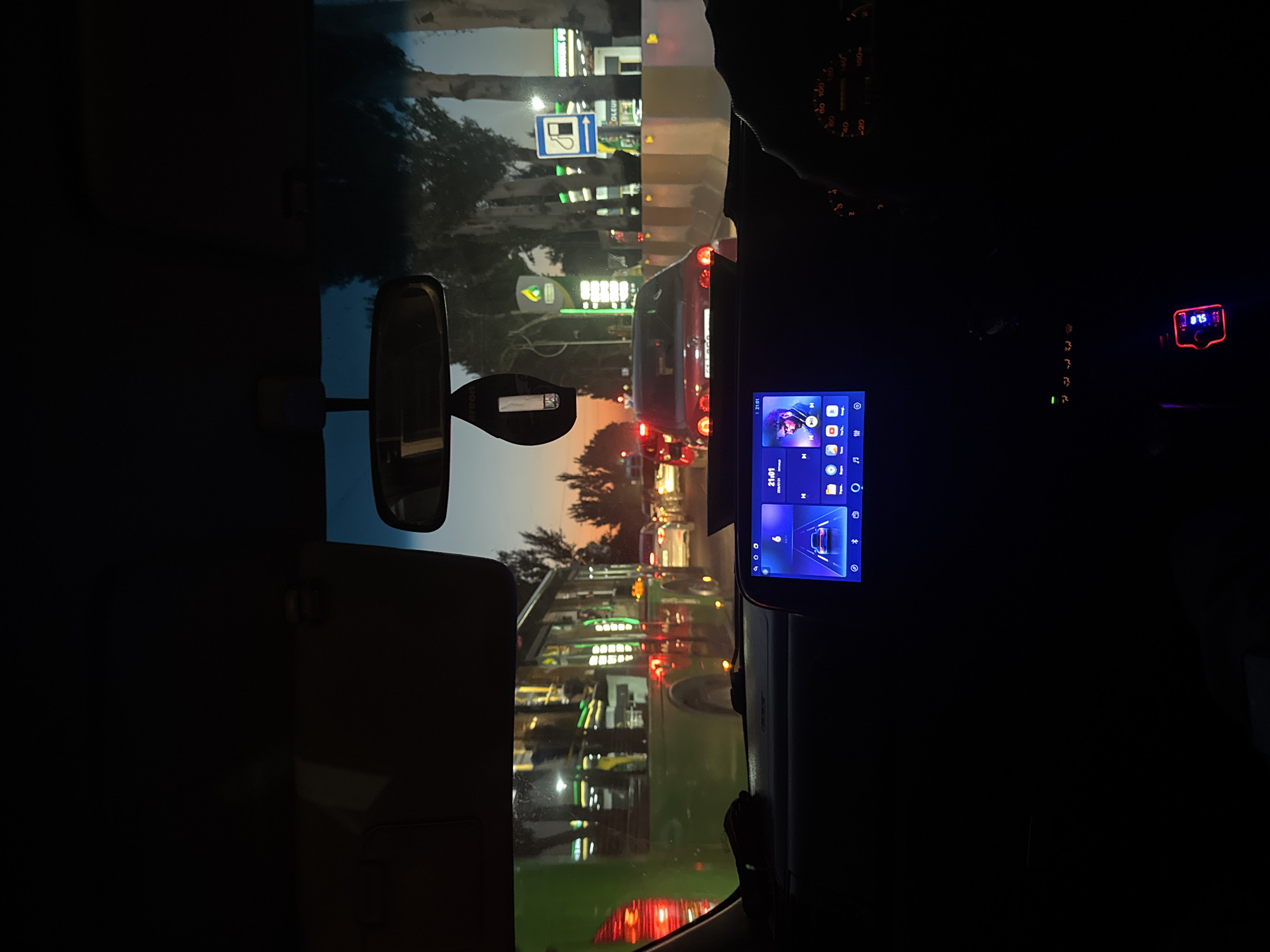 The image depicts a view from inside a car in Bishkek, Kyrgyzstan, during evening traffic. The dashboard features a multimedia screen displaying a car icon, time as 21:01, and music playback information with a picture of a man. To the right of the screen, a portion of the car's speedometer is visible, with the needle pointing between 80 and 100 km/h. A red-lit USB charger is plugged into the car's power outlet in the lower right. Outside the car, traffic is congested, with a green bus in the left lane and several cars ahead. A gas station with illuminated signs is visible on the right side of the road, along with trees lining the street. The sky suggests the sunset is nearing completion.