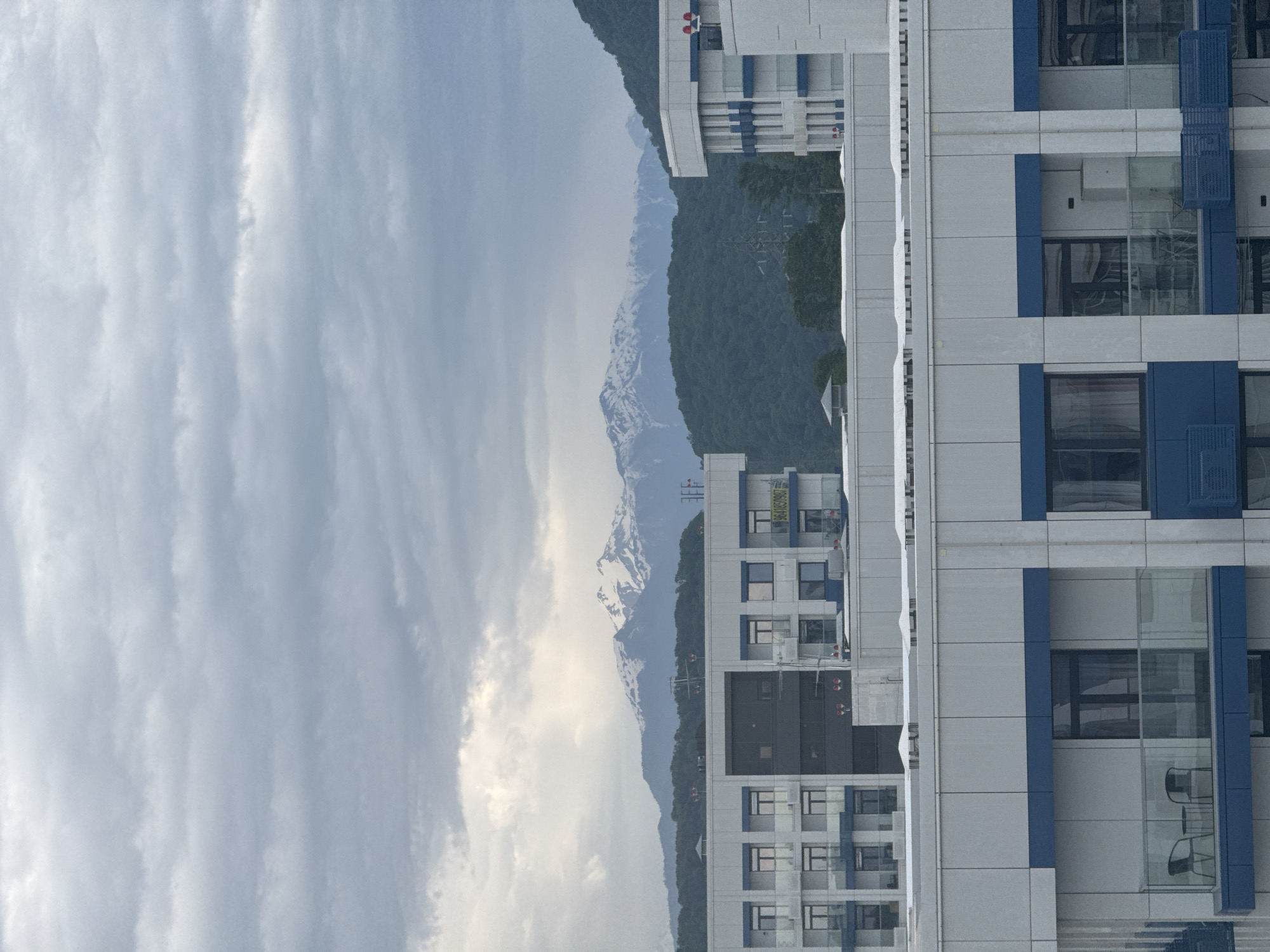 The image depicts a building in Sochi, Russia, set against a backdrop of mountains and cloudy skies. The building is modern, with a gray and blue facade. Some windows feature balconies. The mountains in the background have snow-capped peaks, with forests covering their lower slopes. Power lines run across the mountain landscape. The sky is overcast with heavy, textured clouds.