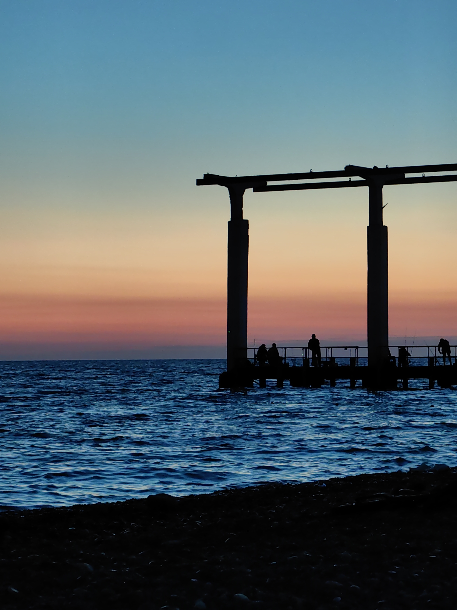 This vertical, silhouette photograph captures a pier extending into the Black Sea at sunset in Sochi, Russia. The sky exhibits a gradient of colors, transitioning from a pale blue at the top to hues of orange, pink, and a faint purple near the horizon. Two large, dark, pillar-like structures, part of the pier's architecture, dominate the upper right portion of the frame. Below these, the pier itself is visible as a dark horizontal line, silhouetted against the colorful sky. Several human figures, also in silhouette, are standing on the pier, some appearing to be fishing with rods. The sea is a deep, textured blue, with visible ripples and waves. In the foreground, a dark, pebbly shoreline completes the scene.