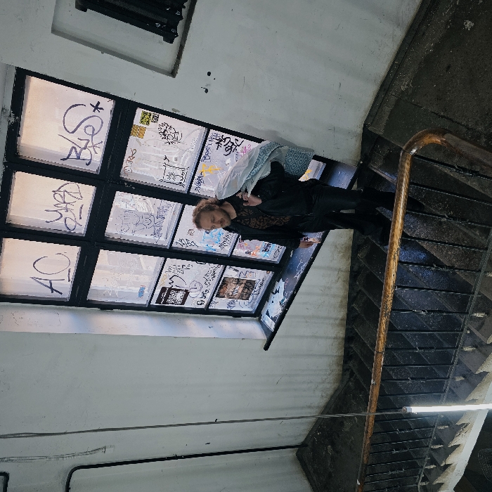 The image depicts an indoor stairwell within an older, multi-story building, consistent with architectural styles found in Saint Petersburg, Russia. The precise location within the city is not discernible. A male individual, appearing in his 30s-40s with light, curly hair and a beard, descends a worn concrete or stone staircase. He wears a dark jacket, a patterned dark shirt, and dark trousers. Over his right shoulder, he carries a light blue and white patterned bundle, resembling a blanket or laundry bag. His gaze is directed towards the lower left. Adjacent to him is a large, multi-paned window with dark frames, heavily obscured by numerous graffiti tags and stickers. Natural light enters through this window. The stairwell features a dark metal railing with a wooden handrail. A vertical fluorescent light fixture illuminates the left side of the scene. Walls are light-colored and show signs of age and wear, with visible electrical conduit on the left.
