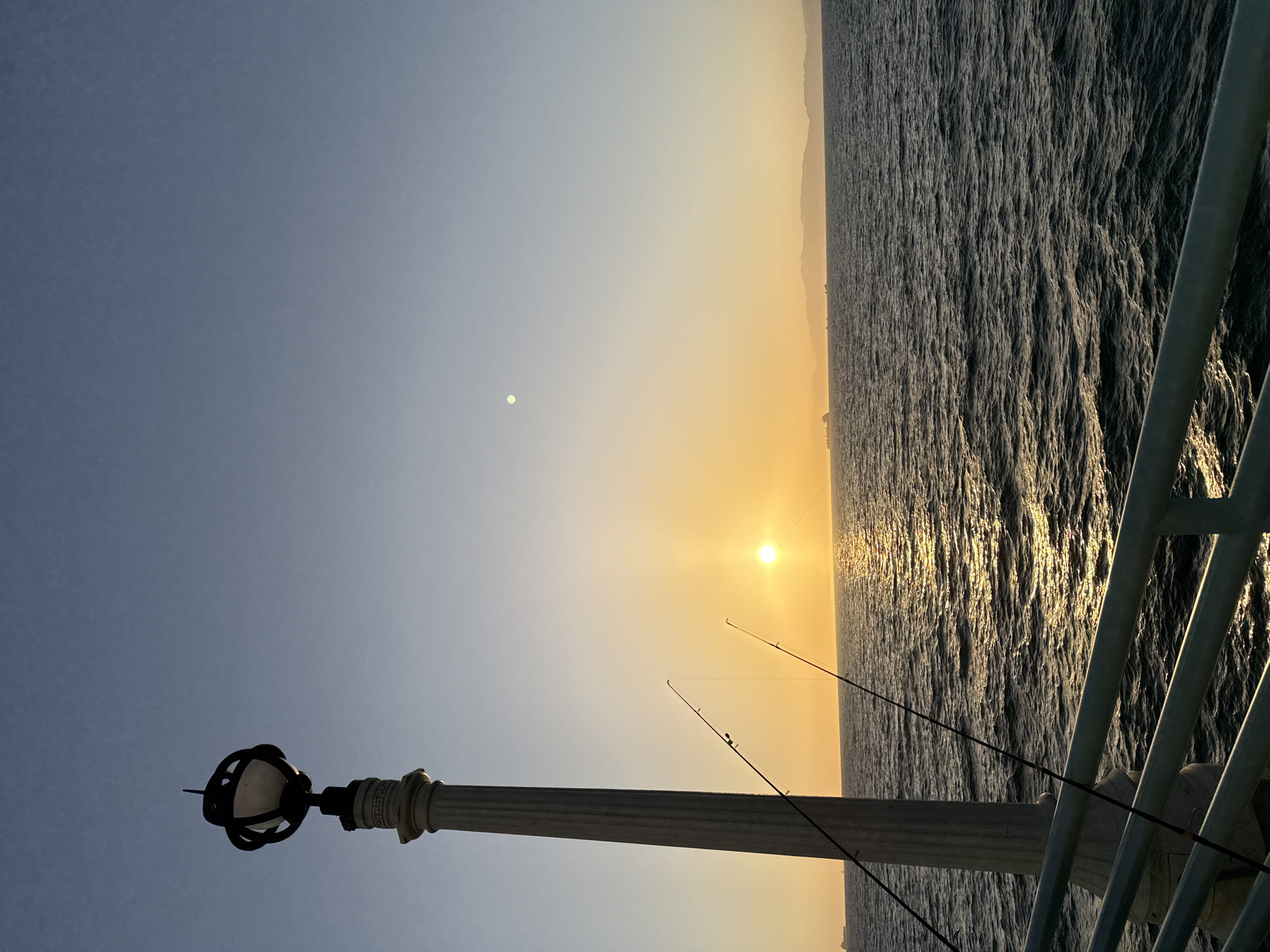 This is a vertical, eye-level, full-shot photograph of a sunset over the ocean from a pier in Manhattan Beach, California. In the foreground, a decorative lamppost with a white column and a black, ornate lamp fixture is positioned on the left side of the frame. Two fishing rods are leaning against the lamppost, with their lines extending towards the water. A pale green railing runs along the bottom of the frame. The ocean fills the middle ground, with moderate waves reflecting the warm hues of the sunset. In the background, a hazy shoreline with hills and mountains is visible, with a few ships or boats dotting the horizon. The sun, a bright orange orb, is near the center of the frame, casting a shimmering golden path across the water. The sky transitions from a pale yellow near the horizon to a clear, soft blue at the top of the frame, with a faint white circle, possibly the moon, visible in the upper portion of the sky. The overall mood is serene and tranquil.