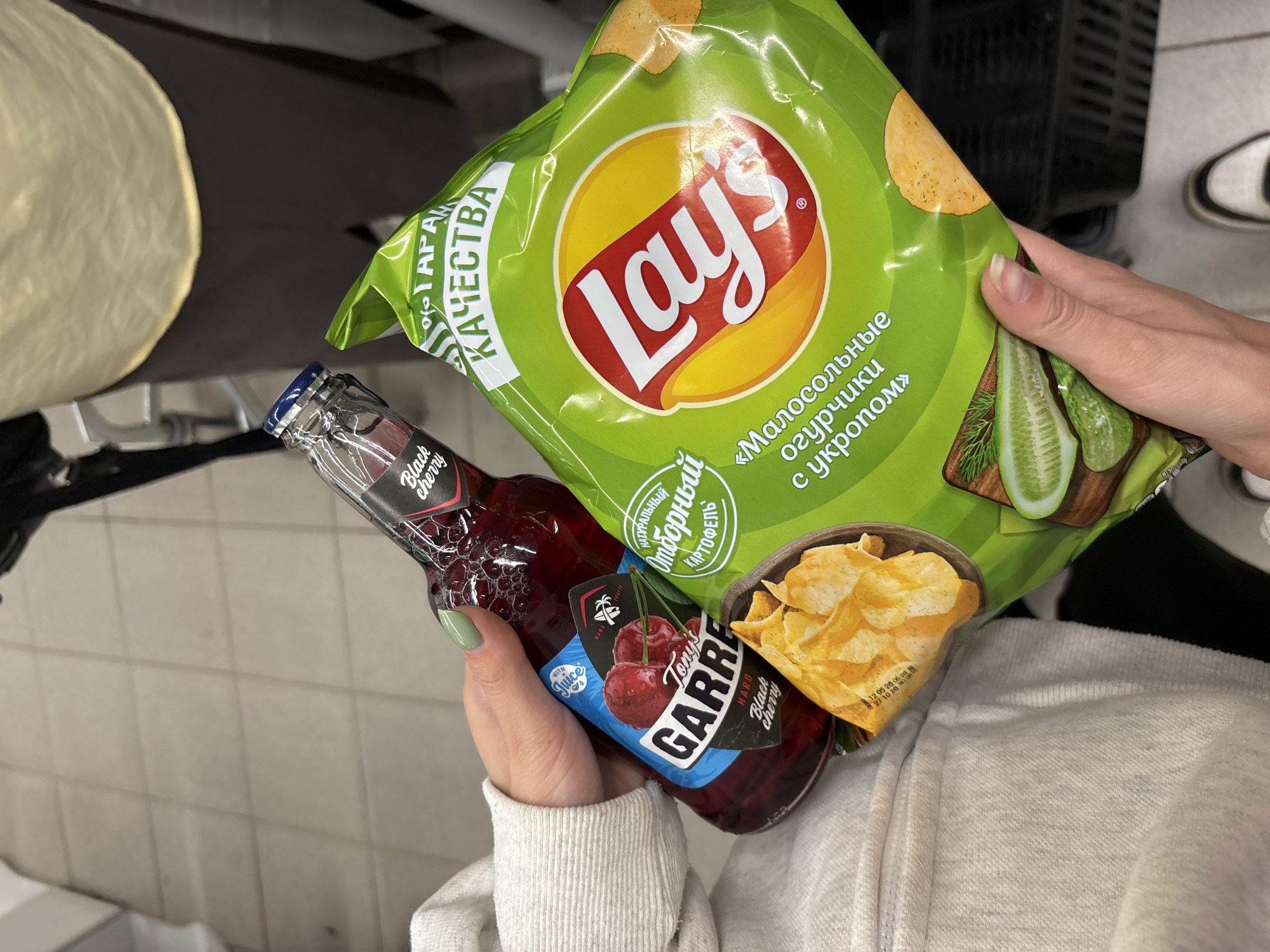 A person's hands are holding a bottle of "Tony's Garre Hard Black Cherry" beverage and a bag of Lay's potato chips. The beverage bottle is made of glass and contains a dark red liquid. The Lay's bag is bright green and features the Lay's logo, along with images of potato chips and dill. The background appears to be a retail environment, possibly a grocery store, with tiled flooring and shopping carts visible. The image seems to have been taken in the city of Yaiva, Russia, indicated by text on the Lay's bag which includes Russian language.