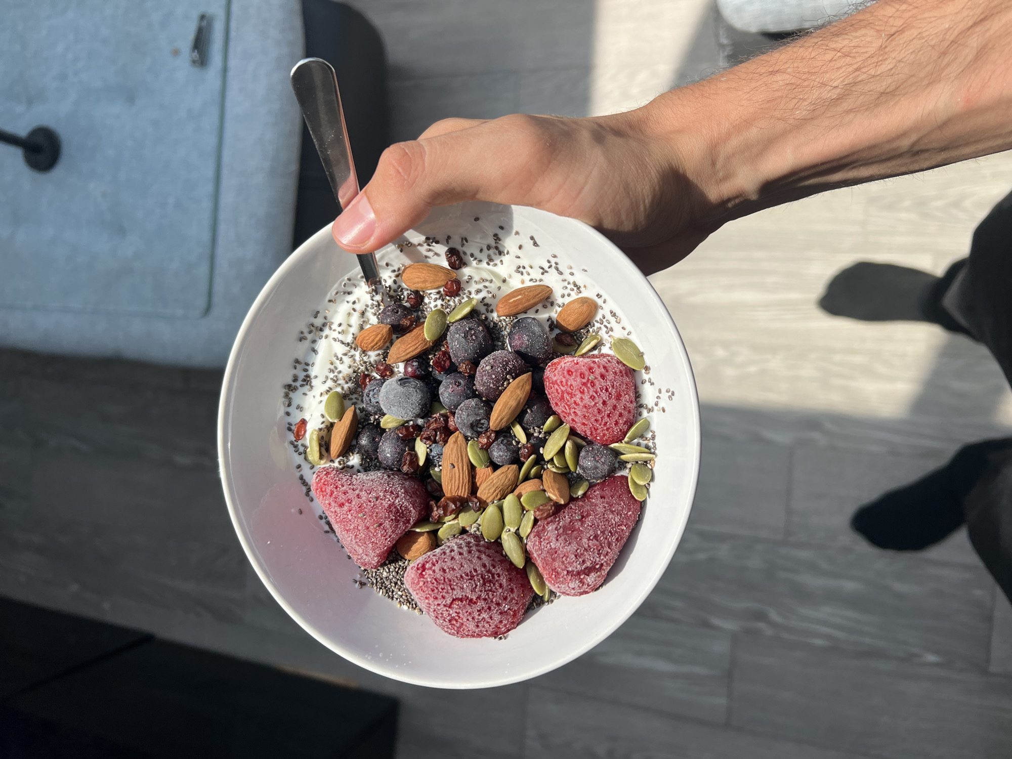 In Rotheux-Rimière, Belgium, a person holds a white bowl filled with yogurt, topped with frozen strawberries, blueberries, almonds, pumpkin seeds, chia seeds, and goji berries. A spoon is partially submerged in the bowl. The background reveals a light-colored surface with subtle patterns, possibly a tabletop, and a muted blue armchair. Sunlight casts a shadow of the person and bowl onto the floor, which appears to be tiled.