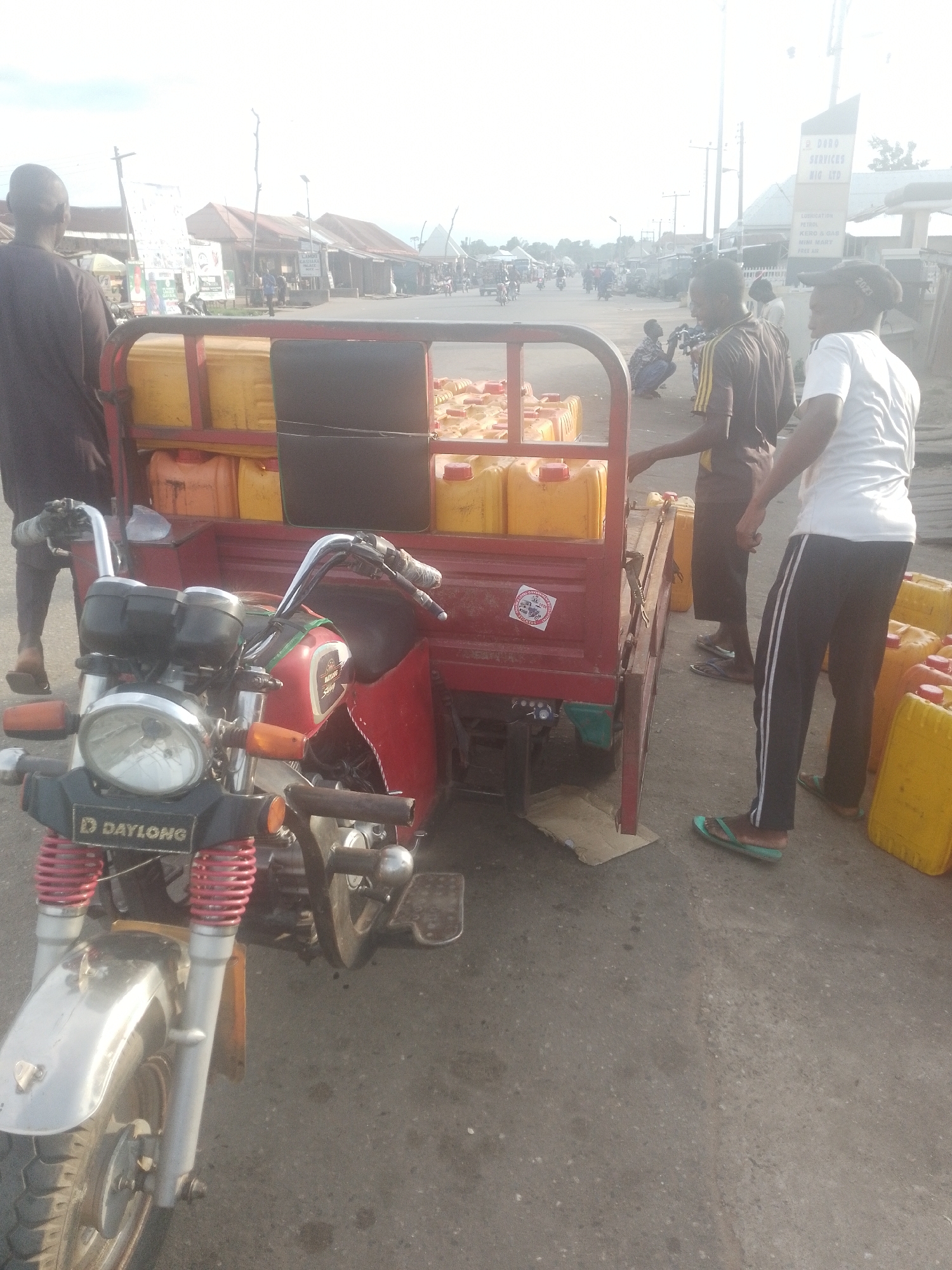 The image depicts a roadside scene in Serti, Nigeria, likely a commercial area given the presence of shops and a busy road. In the foreground, a red three-wheeled motorcycle, possibly a cargo tricycle, is parked. The motorcycle has a prominent "DAYLONG" logo on its front. It is loaded with many large yellow jerrycans, suggesting it is used for transporting liquids, possibly fuel or water.
Several people are visible in the image. A person wearing a dark garment stands to the left, facing away from the camera. To the right of the motorcycle, two young men are standing near the jerrycans. One is wearing a brown and yellow striped shirt and dark pants, and the other is wearing a white t-shirt and black pants with white stripes. Both are wearing flip-flops. Another person is visible in the background, kneeling near a shop.
The background reveals a street with several buildings, some appearing to be shops or stalls. A prominent signpost with text that is partially readable is visible, with "DODO SERVICES NIG LTD" being the clearest part. Further down the street, there is traffic, including motorcycles and other vehicles, indicating an active urban environment. The sky is bright, suggesting it is daytime, possibly a sunny or overcast day. The overall scene conveys a sense of commerce and daily life in a Nigerian town.