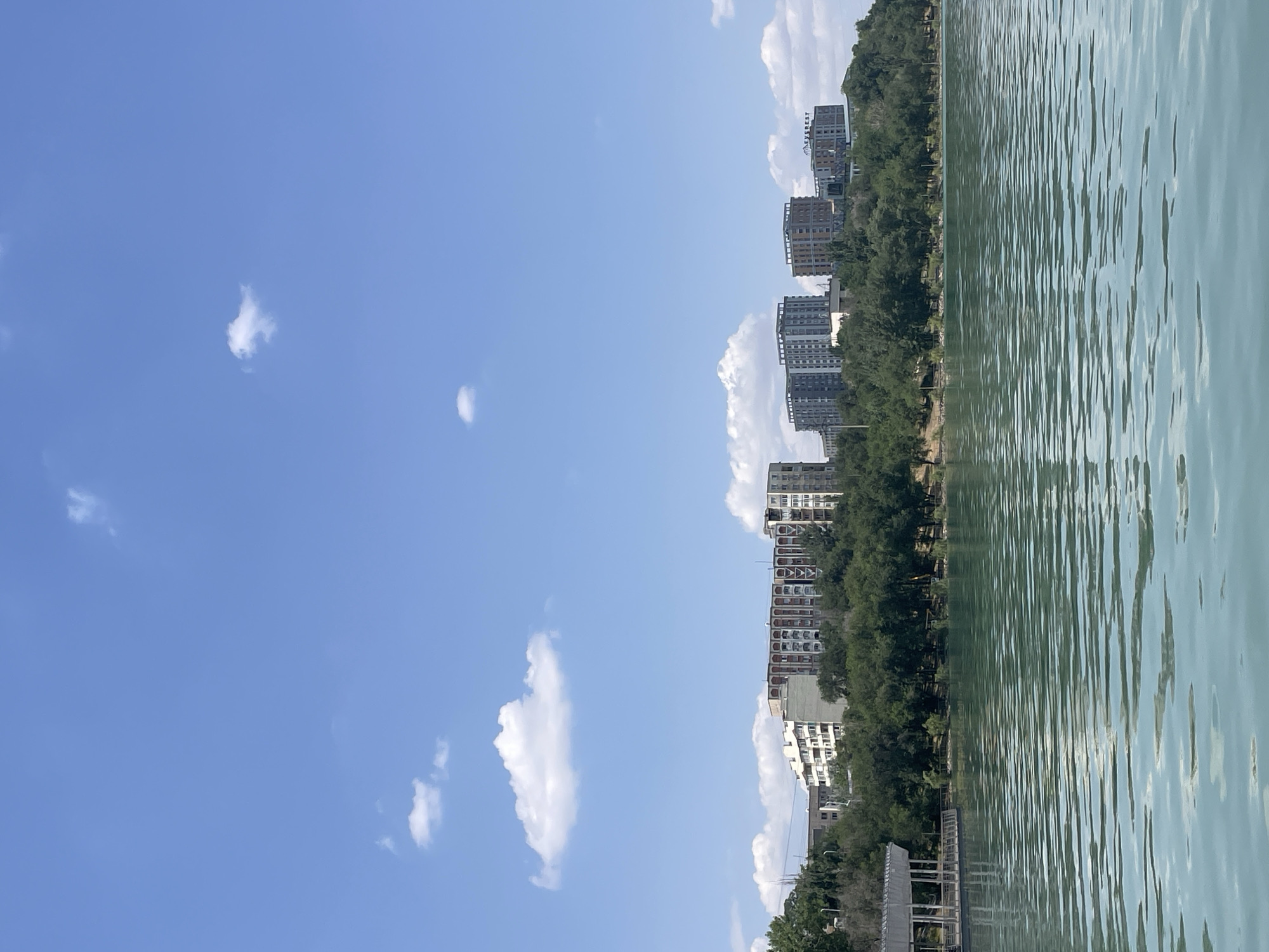 This is a daytime, eye-level shot of a body of water with a city skyline in the background, likely taken from a boat or the shore. The water is a tranquil green and reflects the blue sky, with subtle ripples disturbing the surface. A white gazebo structure with a pier is visible on the left side of the frame, extending over the water.
The background is dominated by a lush green line of trees that separates the water from a collection of modern buildings. These buildings vary in height and architectural style, with some appearing to be residential apartment complexes. One building prominently displays the word "EVEREST" in large letters. The sky above is a clear, bright blue, dotted with a few small, fluffy white clouds.
The scene evokes a sense of urban leisure and modern development coexisting with natural elements. The overall atmosphere is peaceful and serene. The specific location is identified as Almaty, Kazakhstan, suggesting this is a popular recreational area within the city, possibly a park or a waterfront development. There are no people visible in the frame.