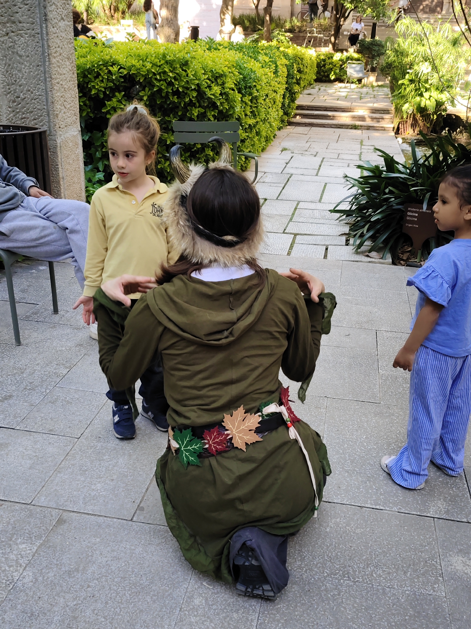 In an outdoor setting, two children are standing on a paved area, one in a yellow shirt and the other in a blue shirt with striped pants. An adult figure, with their back to the viewer, is kneeling and adjusting the child in the yellow shirt's clothing. The adult is wearing a green robe-like garment with a decorative belt adorned with colorful leaf-shaped ornaments, and a headpiece with horns. The environment includes greenery and foliage, indicating a garden or park area. The image was taken in Barcelona, Spain.