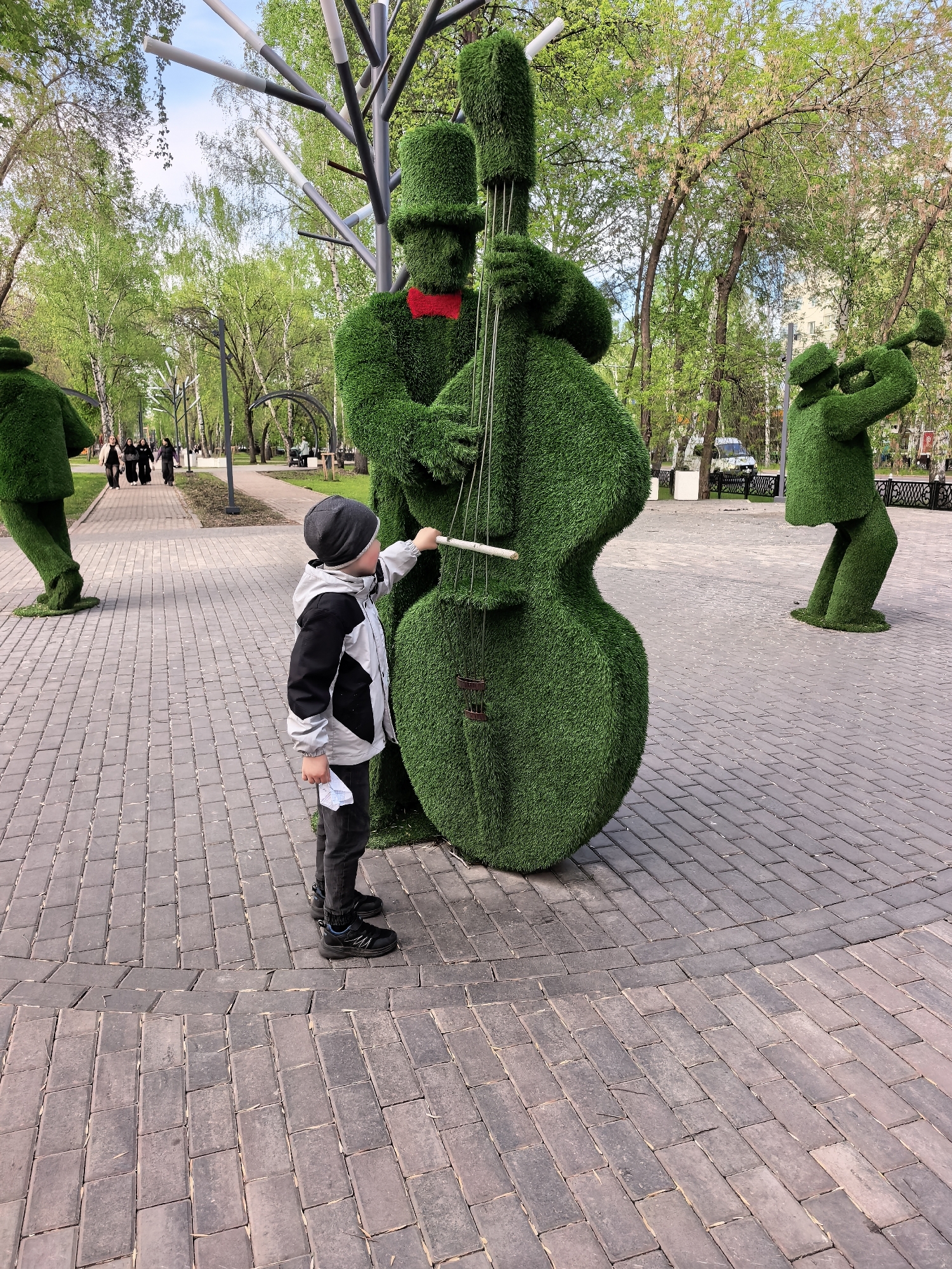 The image depicts a scene in a park in Sterlitamak, Russia, on a brick-paved area. In the foreground, a young boy wearing a grey and black jacket, grey pants, a dark grey beanie, and black sneakers stands next to a topiary sculpture of a musician playing a double bass. The boy is holding a thin white stick and appears to be "playing" the instrument along with the sculpture. The topiary sculpture is made entirely of green material. In the background are other topiary sculptures of musicians, one playing a trumpet and another in a walking pose. In the far background, there is a park with green trees and a path with people walking. The sky is visible through the trees. A black metal fence can be seen behind the musician with the trumpet.