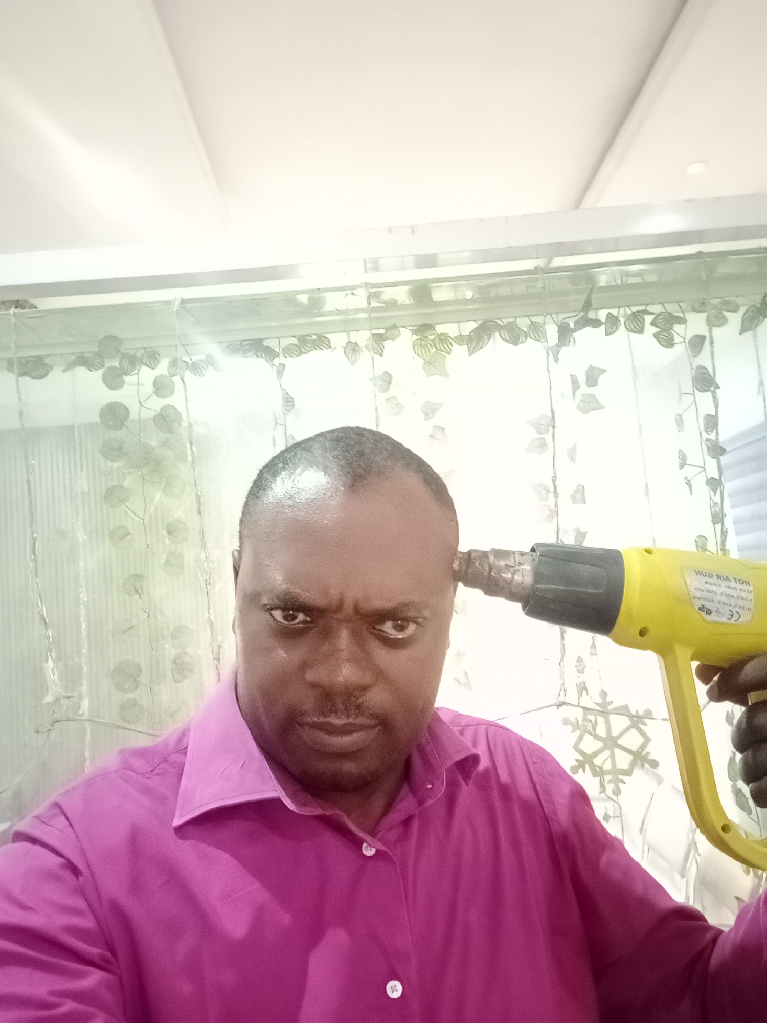A man is holding a yellow heat gun to his head. He is in front of a window with translucent curtains. The man is wearing a pink shirt. The location is likely a residence, given the curtains and interior setting. There is no explicit information indicating the specific location in Gwarinpa, Nigeria.