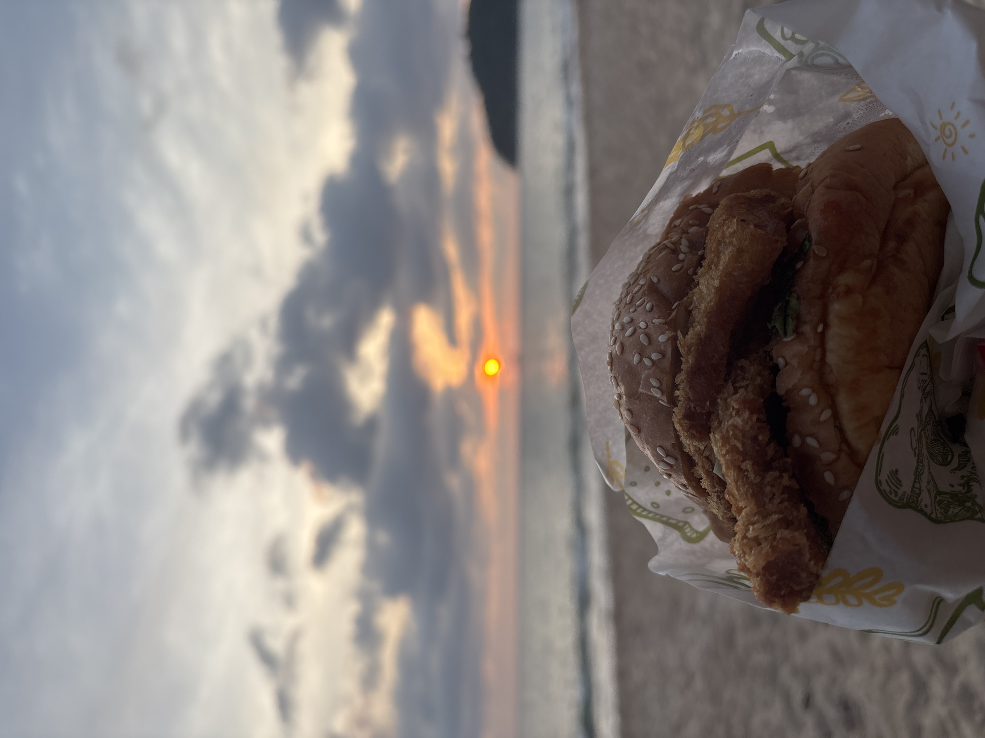 The image captures a close-up of a burger held in a branded paper wrapper, set against a blurred backdrop of a beach sunset. The burger, positioned on the right, features a sesame seed bun and a thick, breaded, fried patty (likely chicken or fish), with hints of green lettuce visible.
The background reveals a serene sunset over the ocean. A distinct, bright orange-yellow sun is visible low in the sky, casting warm light across diffused clouds and the water. The sky displays a mix of darker grey clouds and lighter, illuminated areas, characteristic of a setting sun. The sandy beach is partially visible in the lower right, suggesting a coastal location. The image is composed with a shallow depth of field, rendering the burger in sharp focus while the sunset scene remains softly blurred.
The wrapping paper around the burger is white with stylized green and yellow patterns, including a clear yellow sun icon with rays. The time of day is clearly sunset or late golden hour, indicated by the sun's position and the warm, soft lighting. The overall scene evokes a relaxed moment at the beach in Choeng Thale, Thailand. No specific textual words are clearly legible on the wrapper.