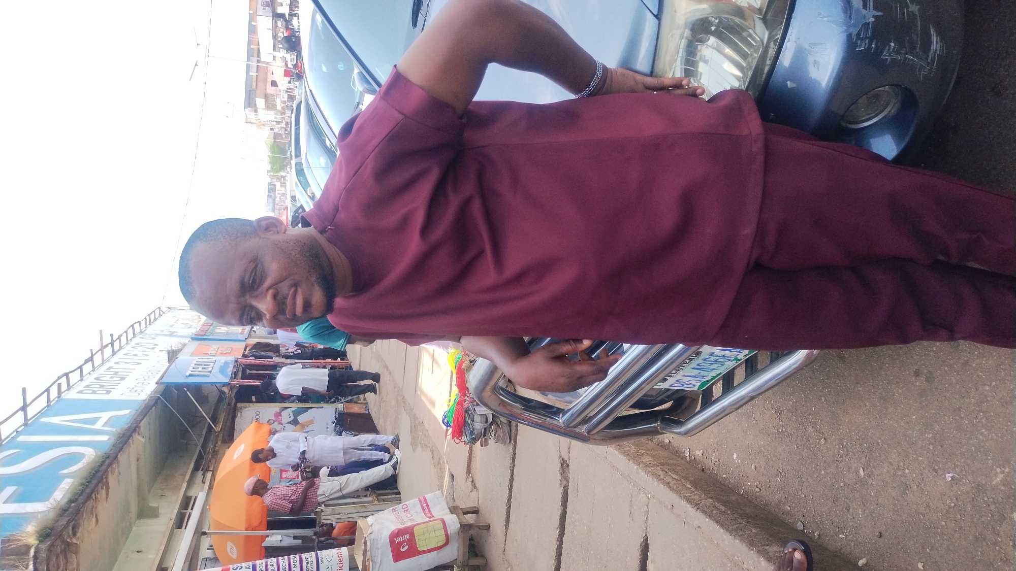 A man in a maroon traditional outfit leans against a dark blue car. He has a short beard and is looking at the camera with a slight smile. The car has a chrome bumper and a license plate that reads "PLATEAU" and "BG4A96E". The scene appears to be outdoors, possibly in a market or along a street, given the presence of various stalls and people in the background.
Behind the man, a busy street scene unfolds. A large blue sign with white lettering that partially reads "ESA" and "BRIGHT 3/6/20 CB" is visible. Further down, other signs and stalls are present, including one with an orange umbrella. Several people, some in white attire, are seen walking or standing around. There are also some items for sale, such as what appears to be SIM cards and cables. The overall atmosphere suggests a bustling commercial area. The lighting indicates it is daytime, with no clear indicators of weather. The presence of "PLATEAU" on the license plate and the signs in English point to Nigeria as the location.