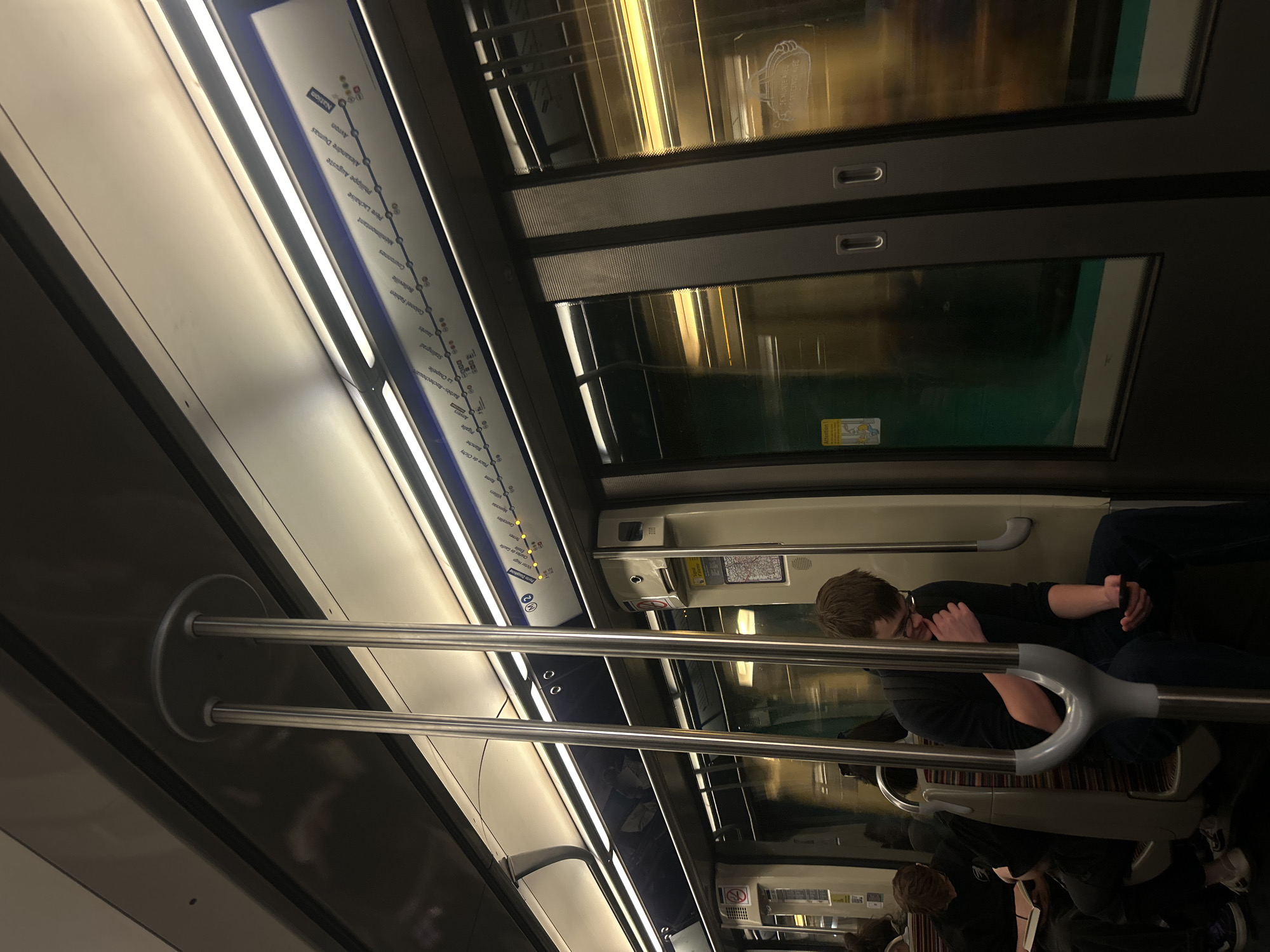 This photo shows the inside of a subway car, likely in Paris, France. A subway route map is affixed to the wall above the passengers. The stations indicated on the route are Nation, Avron, Alexandre Dumas, Philippe Auguste, Père Lachaise, Ménilmontant, Couronnes, Belleville, Colonel Fabien, Jaurès, Stalingrad, La Chapelle, Barbès-Rochechouart, and Gare du Nord. A person is seated in the subway car, holding a smartphone. There are two horizontal bars for standing passengers. The interior is brightly lit. Other passengers are visible in the reflection of the subway car windows.