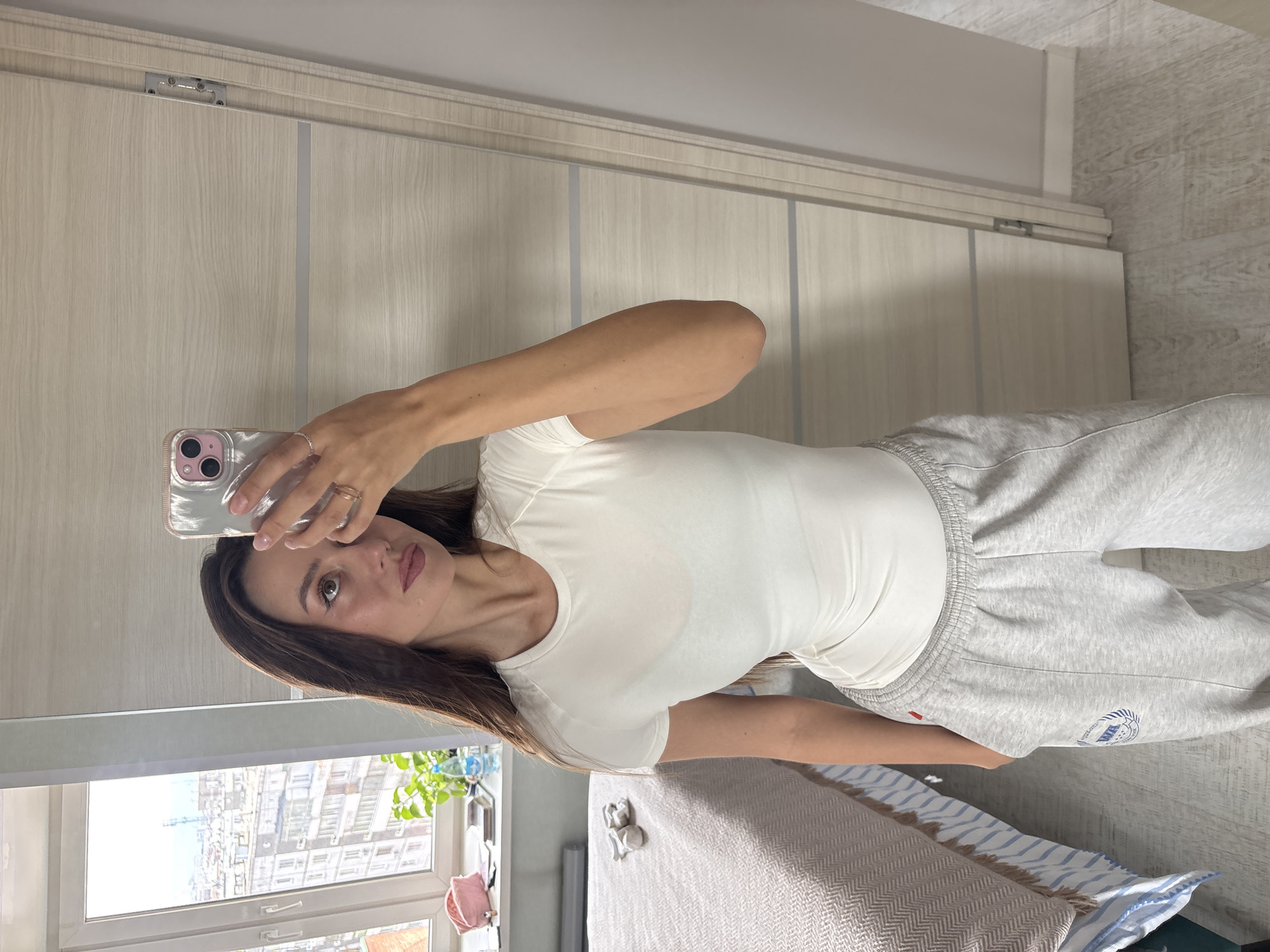 A brunette woman in a white t-shirt and gray sweatpants takes a selfie with her smartphone. She is indoors with a window behind her showing a cityscape with buildings, potentially the city of Krasnoyarsk, Russia. A bed is partially visible in the lower right of the frame. Behind her is a closet with light wood-grain doors.