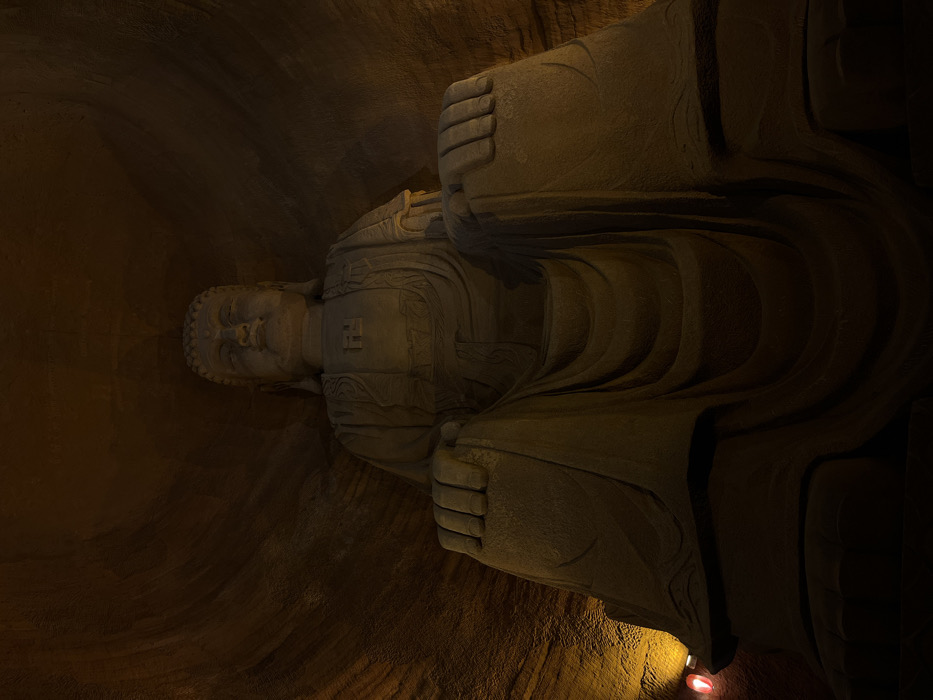 The image displays a monumental, light-colored stone Buddha statue carved into a dark rock formation within a cavern, located in Shizhong, China. The statue is depicted in a seated, meditative posture with hands resting on its lap, occupying the central and upper portions of the frame when viewed from a low-angle perspective. A prominent clockwise swastika symbol (卍) is carved onto the chest of the figure. The surrounding cavern walls, from which the statue appears to be partially carved, exhibit horizontal striations and a rough, natural texture. The scene is illuminated by warm, localized light sources from the lower left, casting areas of the statue and rock in yellow-orange light while leaving upper sections and deep crevices in shadow. A small red-glowing object and a distinct yellow light source are visible at the base of the left cavern wall. No human subjects are present apart from the statue.