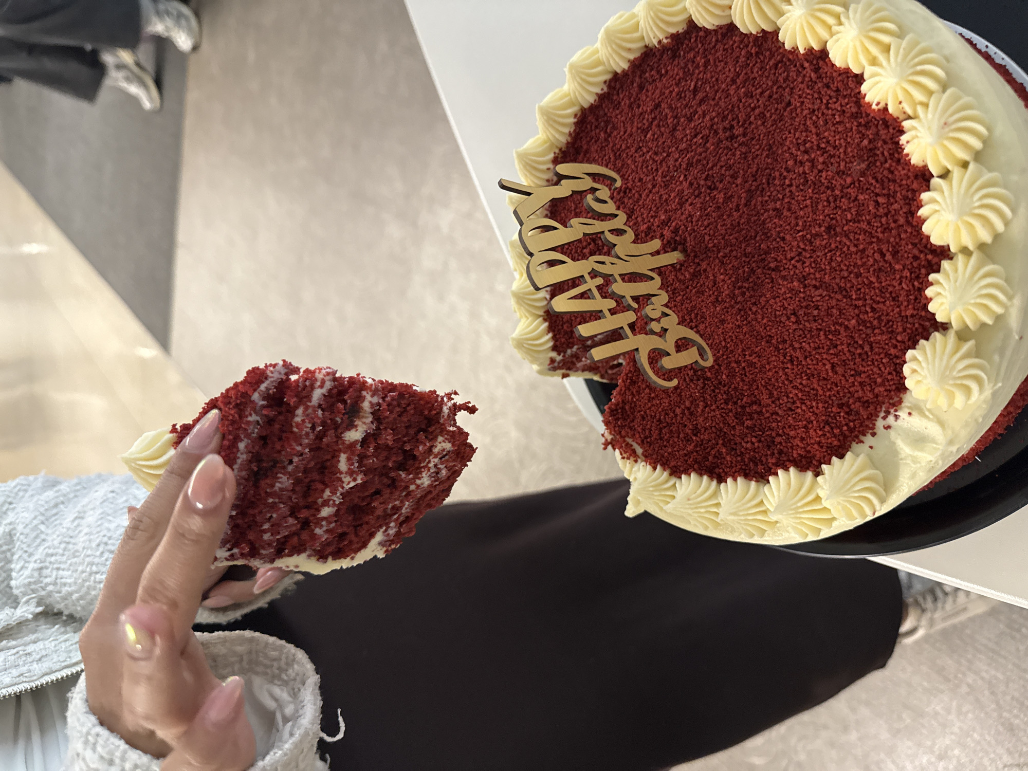A partial view from above shows a red velvet cake and a slice of the cake held in a hand. The cake is round with white frosting rosettes around the perimeter. A "Happy Birthday" cake topper is inserted into the cake where a slice has been removed. The cake sits on a black plate. A hand with manicured nails holds a piece of the cake, also frosted with white frosting. The person appears to be sitting, wearing a black garment with a white jacket or sweater. The background is a beige surface. The location is in Mexico City, Mexico.