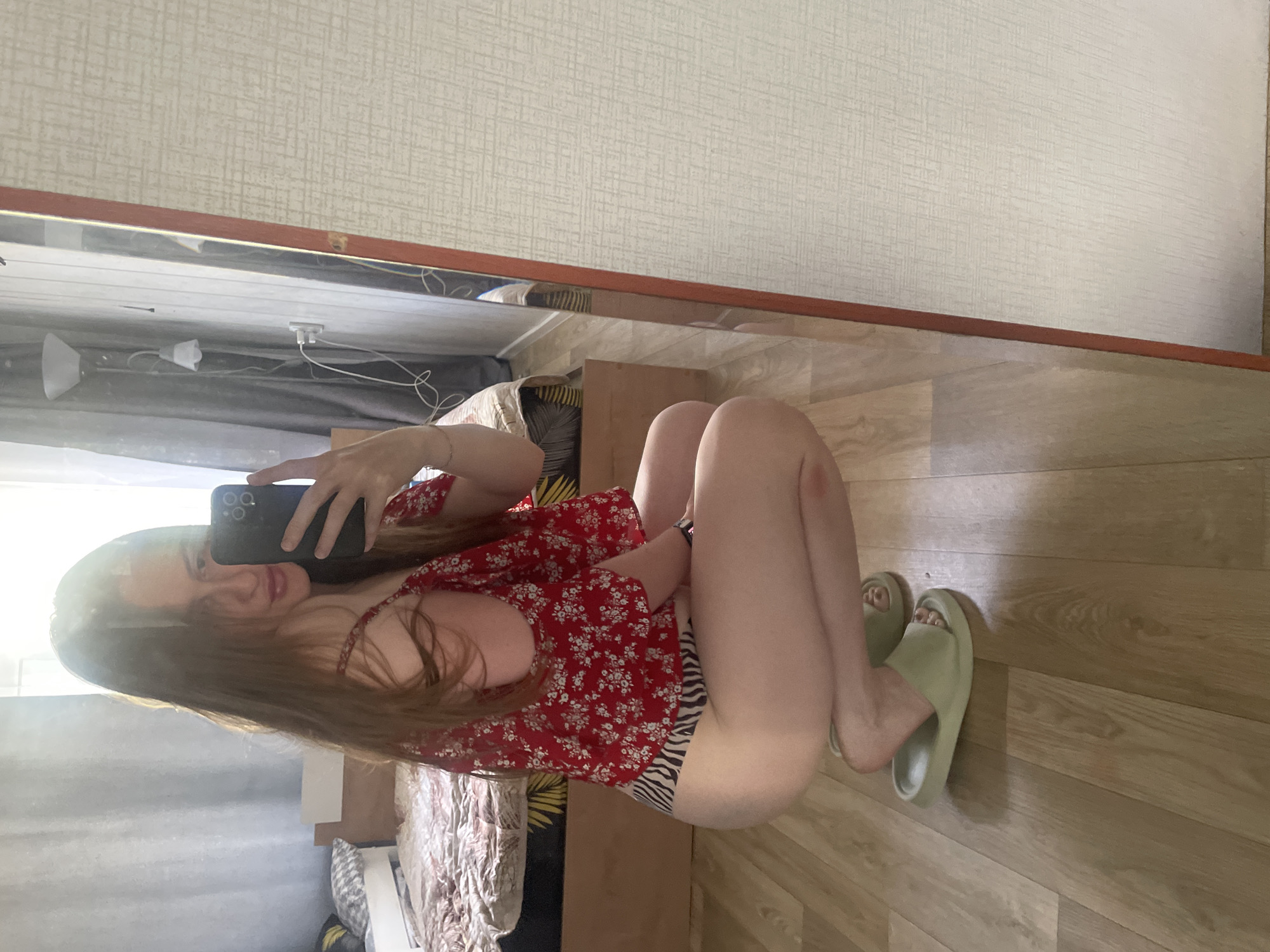 A young woman with long, auburn hair is taking a selfie in a bedroom. She is wearing a red, floral print top and zebra-striped underwear. She is squatting on a light brown wooden floor, facing a large mirror. The room appears to be a bedroom, with a bed visible in the background, covered with a patterned comforter. Curtains hang by a window. A floor lamp is also visible. The overall lighting suggests it is daytime. The image is cropped to focus on the woman and her reflection in the mirror. The location is specified as Syrokovo, Russia.