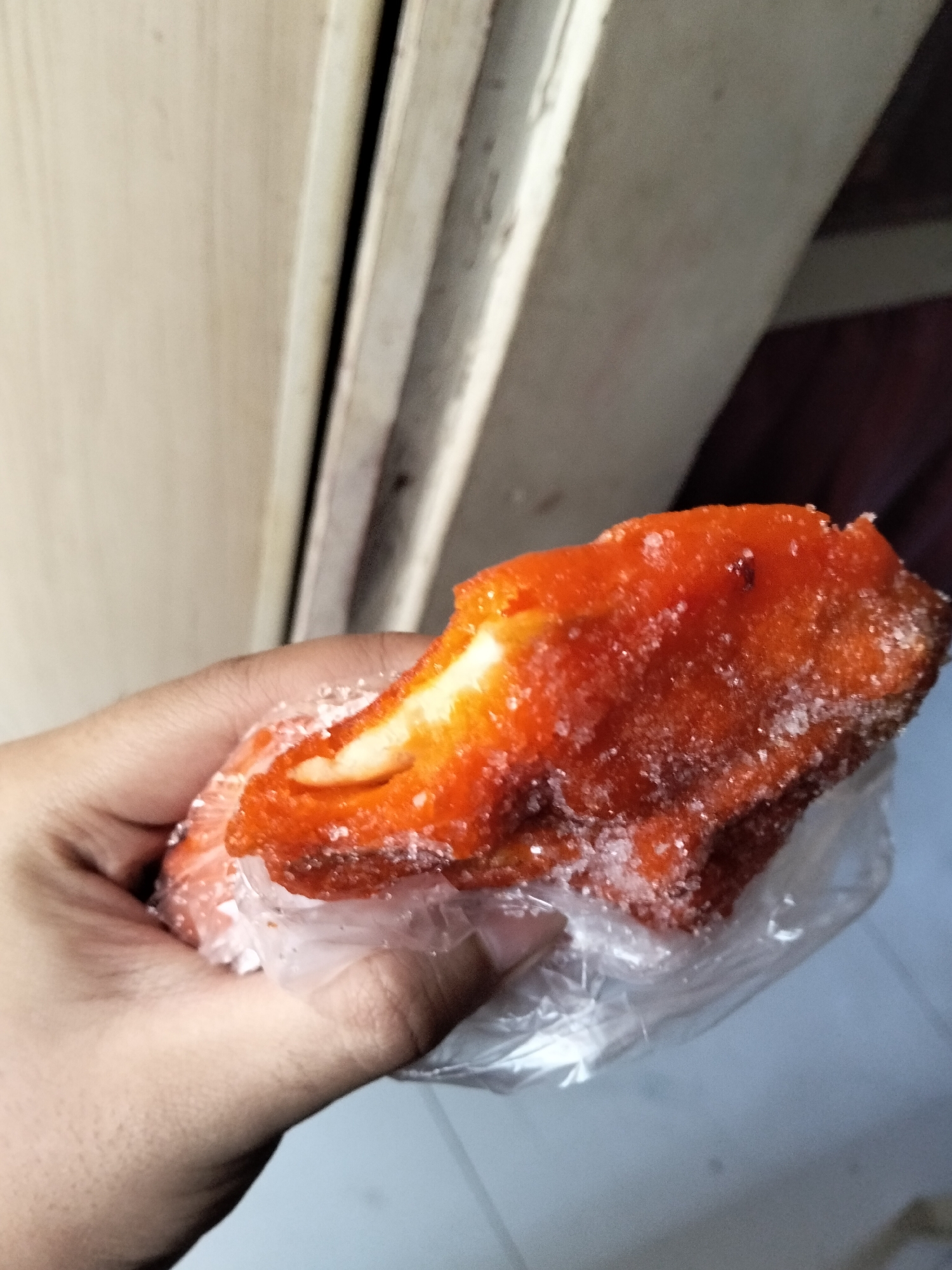A hand is holding a sweet pastry, possibly a donut or a fritter, coated in orange sugar and sugar crystals. The pastry has a light-colored filling visible on one side. It is partially wrapped in clear plastic. The background shows a beige wall with vertical paneling on the left and a darker, out-of-focus area on the right. The lighting suggests an indoor setting. The scene is not definitively located in Davao City, Philippines, as there are no recognizable landmarks or contextual clues to confirm this specific location.