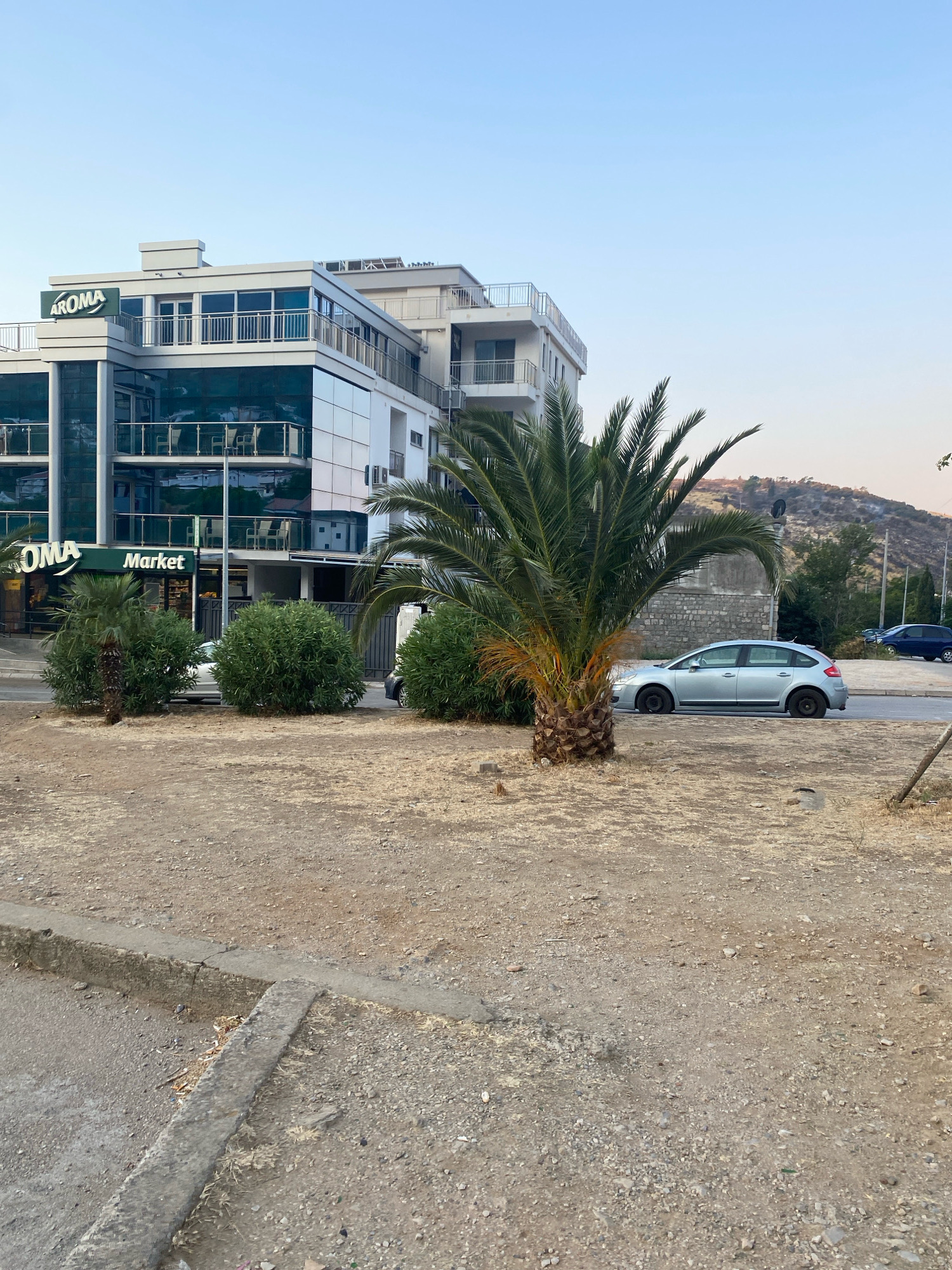 The image depicts a street scene in Sutomore, Montenegro, featuring a modern, multi-story building with a prominent "AROMA Market" sign. The building has a glass facade on its lower levels and balconies on the upper floors. In front of the building, a silver hatchback car is parked on the side of the road. A palm tree stands near the car, with its fronds extending over the roadway. The foreground of the image is a dry, gravelly area with a concrete curb. In the background, a hilly landscape is visible, with some sparse vegetation and a clear blue sky. The overall impression is of a commercial area in a Mediterranean climate.