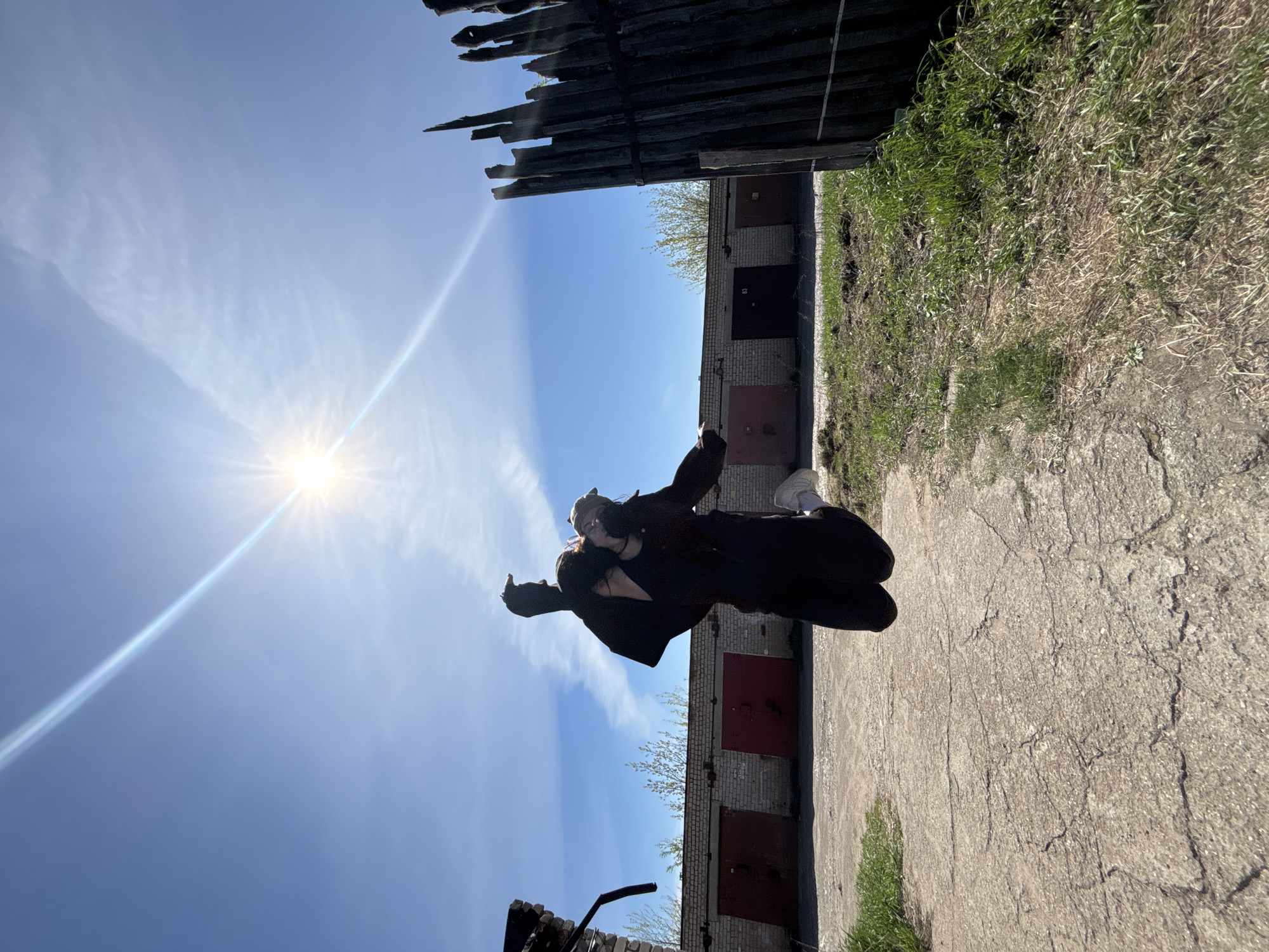 An individual is posing in an outdoor scene. The person, wearing a dark outfit and light-colored sneakers, is positioned between a weathered wooden fence and a row of brick garages with metal doors. The background features a clear blue sky with the sun shining brightly. The ground is a cracked concrete surface, and there is some grass visible along the edges of the path. The city of Perm, Russia is not identifiable from this image.