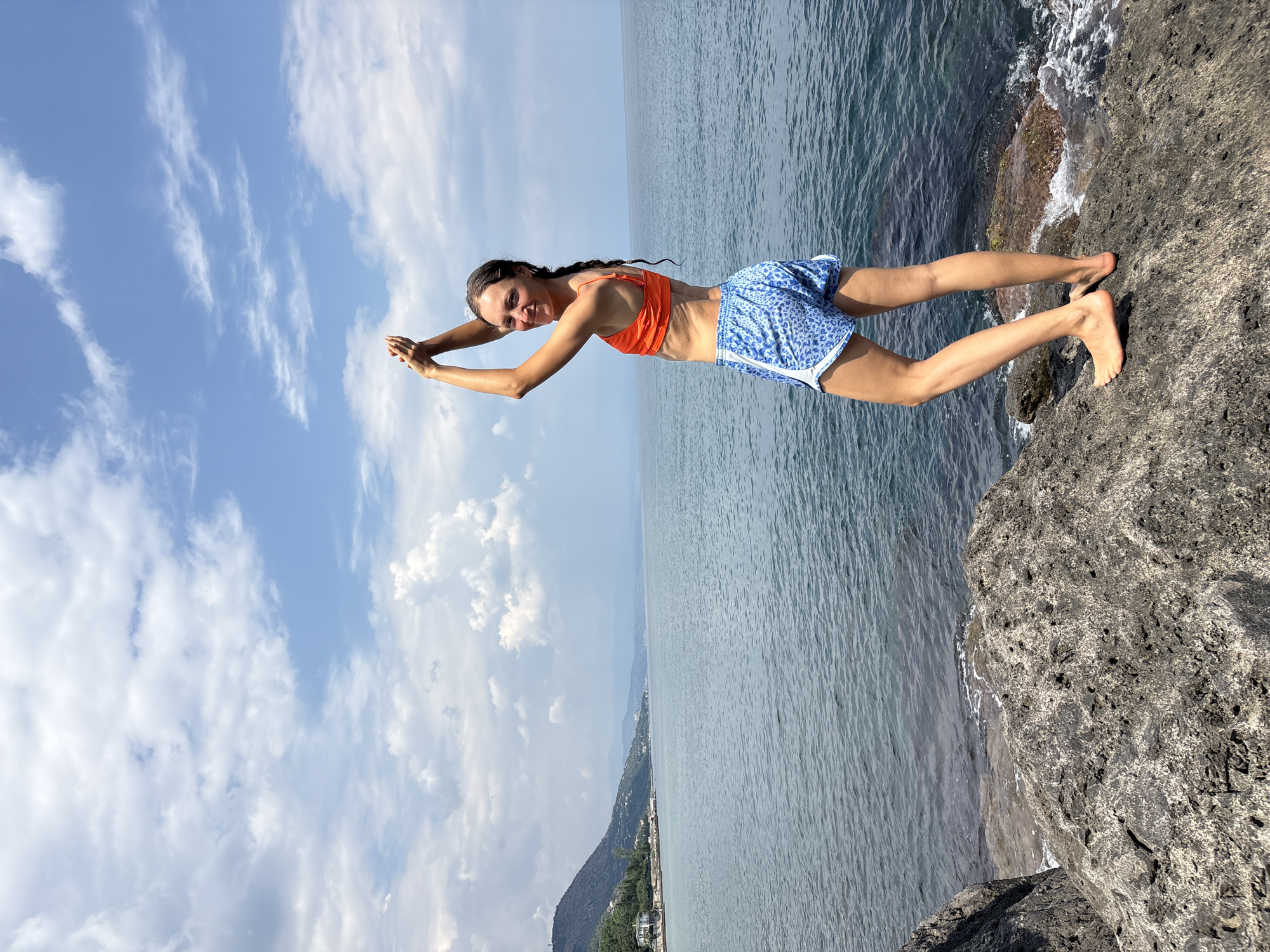 A woman with dark hair in a braid stands on rocks by the sea. She is wearing an orange bikini top and blue and white patterned shorts. Her arms are raised above her head, and her hands are clasped. She is looking towards the camera and smiling. The sea is calm, and the sky is cloudy. In the background, there is a coastline with trees and buildings. The location is the city of Bondarenkovo, Ukraine.