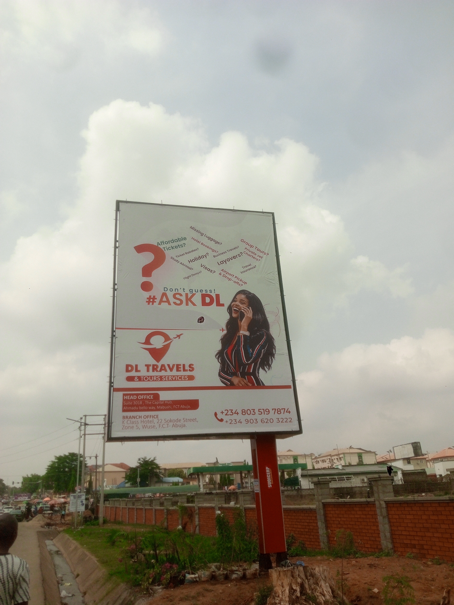 A large billboard for "DL TRAVELS & TOURS SERVICES" stands prominently in an urban setting in Abuja, Nigeria, under a cloudy, overcast sky. The billboard's design features a large red question mark on the left, surrounded by various travel-related questions in white text such as "Affordable Tickets?", "Missing Luggage?", "Hotel Bookings?", "Holiday?", "Study Abroad?", "Group Tours?", "Private Jet Charters?", "Business Travels?", "Layovers?", "Travel Insurance?", "Visas?", "Flight Delays?", and "Airport Pickup & Drop-offs?".
Below the question mark, the slogan "Don't guess! #ASK DL" is displayed. To the right of the billboard is an image of a smiling woman holding a phone to her ear. The company's logo, a red location pin with an airplane icon, is centered below the main graphic, above the company name "DL TRAVELS & TOURS SERVICES."
Visible text includes the "HEAD OFFICE" address: "Suite 3018, The Capital Hub, Ahmadu bello way, Mabushi, FCT-Abuja," and the "BRANCH OFFICE" address: "K Class Hotel, 22 Sokode Street, Zone 5, Wuse, F.C.T-Abuja." Two contact phone numbers are listed: "+234 803 519 7874" and "+234 903 620 3222." The billboard's support pole is red, with the word "SUNDRUP" visible on its side.
The surrounding scene shows a daytime street environment. In the foreground, there's a patch of dirt with a tree stump and some green foliage. An open concrete drainage ditch runs alongside a paved area where the back of a person is partially visible. Further down the street, there are other people, cars, and buildings, including what appears to be a gas station with a green roof. A brick wall separates the immediate foreground from some of the distant structures. The overall impression is of a busy, commercial area.