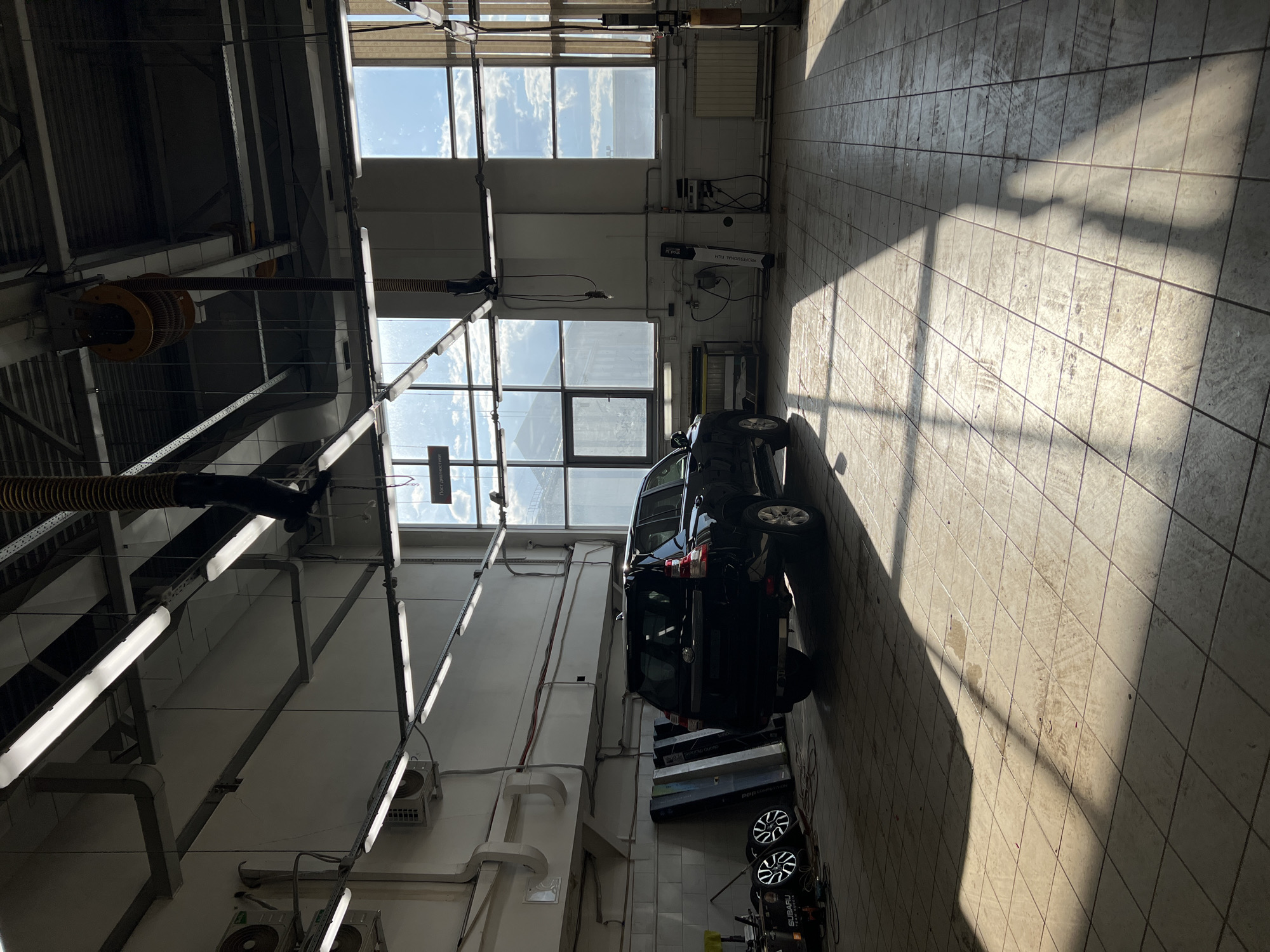 This image depicts the interior of a vehicle service bay located in Izhevsk, Russia. No individuals are present within the frame.
A dark-colored Toyota Land Cruiser Prado SUV is prominently parked in the left-center, with its rear facing towards the camera and its front oriented towards the left wall. The vehicle rests on a light-colored tiled floor that shows signs of wear and minor stains.
The primary light source is natural sunlight entering through a series of large, multi-pane windows that constitute the majority of the right wall. These windows cast distinct, elongated diagonal shadows of their frames and internal structures across the tiled floor. Overhead, numerous industrial-style fluorescent light fixtures are mounted directly to the ceiling or suspended by wires, providing artificial illumination. Flexible yellow and black ventilation hoses descend from the ceiling, with one coiled and another hanging down on the right side.
The left wall features several mounted components, including air conditioning units and exposed conduit pipes. On the floor near the rear of the SUV, a collection of items is arranged: four vehicle wheels with silver multi-spoke rims, several large rectangular panels leaning against the wall, a yellow and black box with "SUBARU TEAM RUSSIA" text, and assorted cables and tools.
Along the right wall, a tall, white cylindrical roll labeled "PROFESSIONAL FILM" stands upright. Below the windows on this side, a radiator is installed. Wall-mounted electrical boxes and equipment are also visible. A sign with the Cyrillic text "Пост диагностики" (Diagnostics Post) is affixed to the upper section of the wall near the windows.
Through the large windows, a bright blue sky with scattered white clouds is visible, alongside glimpses of distant buildings and structures, indicating an urban or industrial exterior. The overall environment is functional and industrial, characteristic of an automotive service facility.