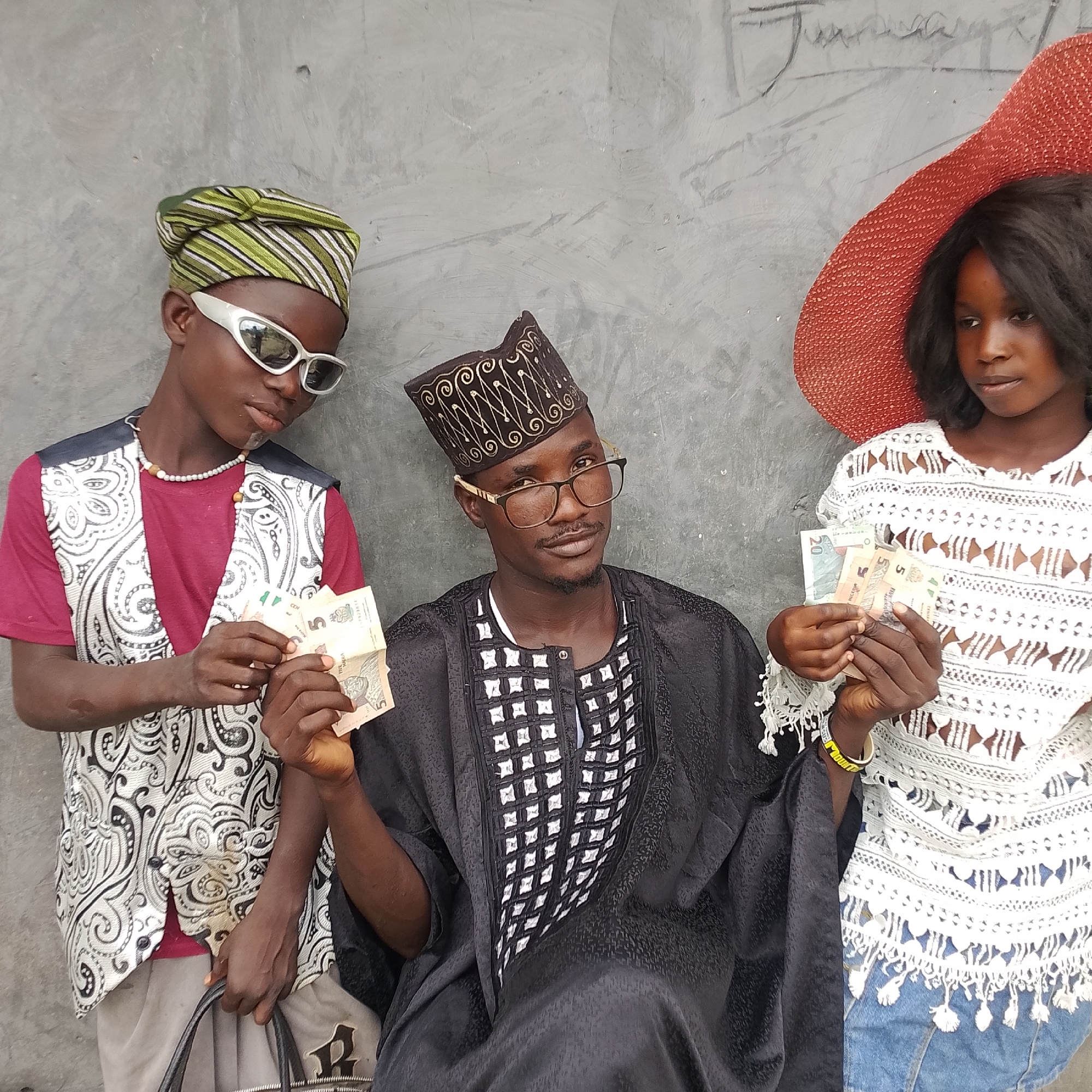 In Ado, Nigeria, three individuals stand against a textured grey wall. On the left, a person wears a green and white striped headwrap, white sunglasses, a burgundy t-shirt, and a white and black patterned vest, holding several banknotes. In the center, a person in a black patterned cap and a black traditional robe with white detailing, wearing glasses, looks directly at the camera, also holding banknotes. On the right, a person in a large red hat and a white lacy top, with dark hair, holds banknotes and looks to their left. The wall behind them is rough and uneven, with some faint markings at the top. The scene appears to be outdoors, possibly in an urban setting, given the wall texture.
